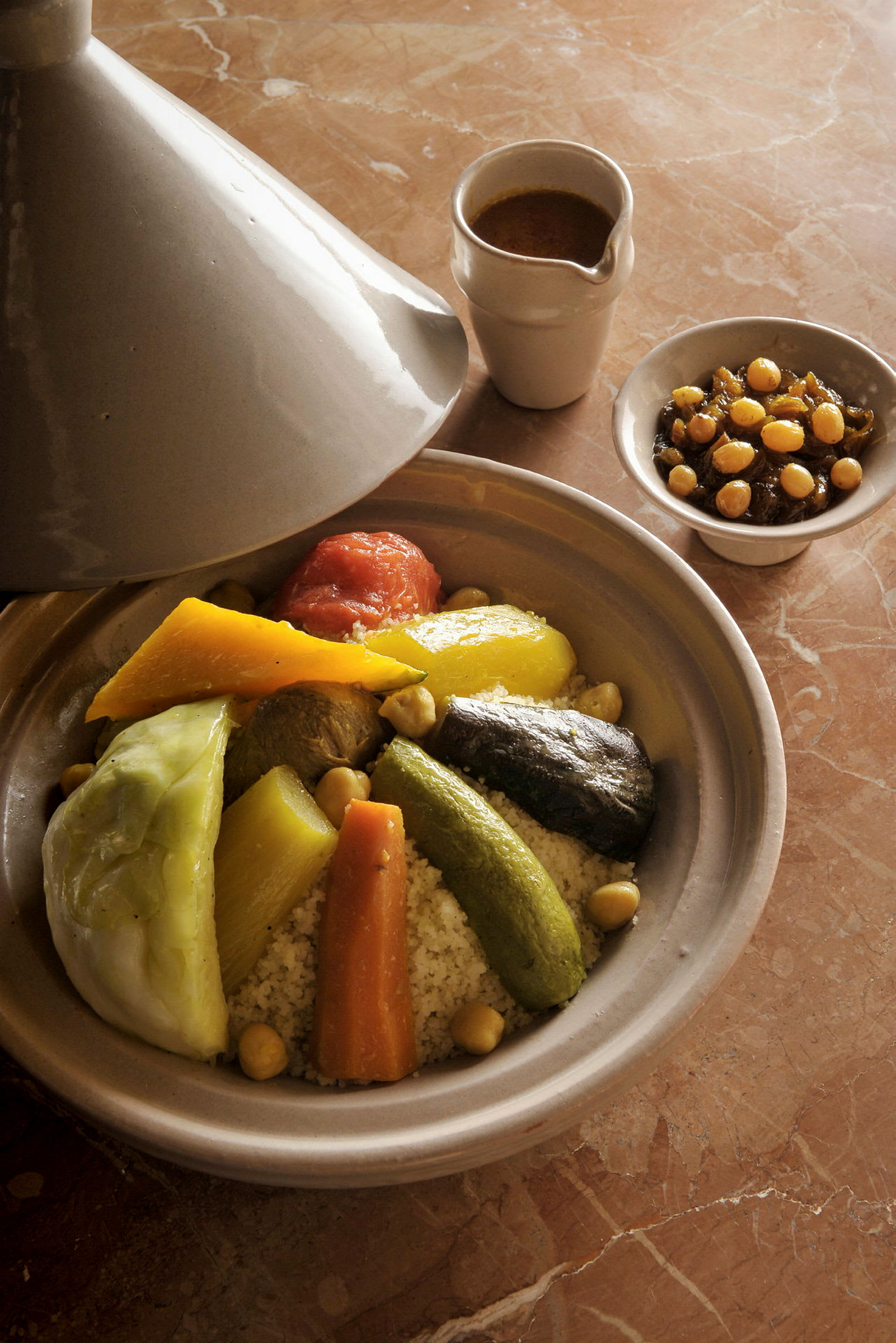 En traditionel tagine-gryde med grøntsager og couscous, serveret med sauce og en skål med kikærter. På et marmorbord. (AI-genereret tekst)