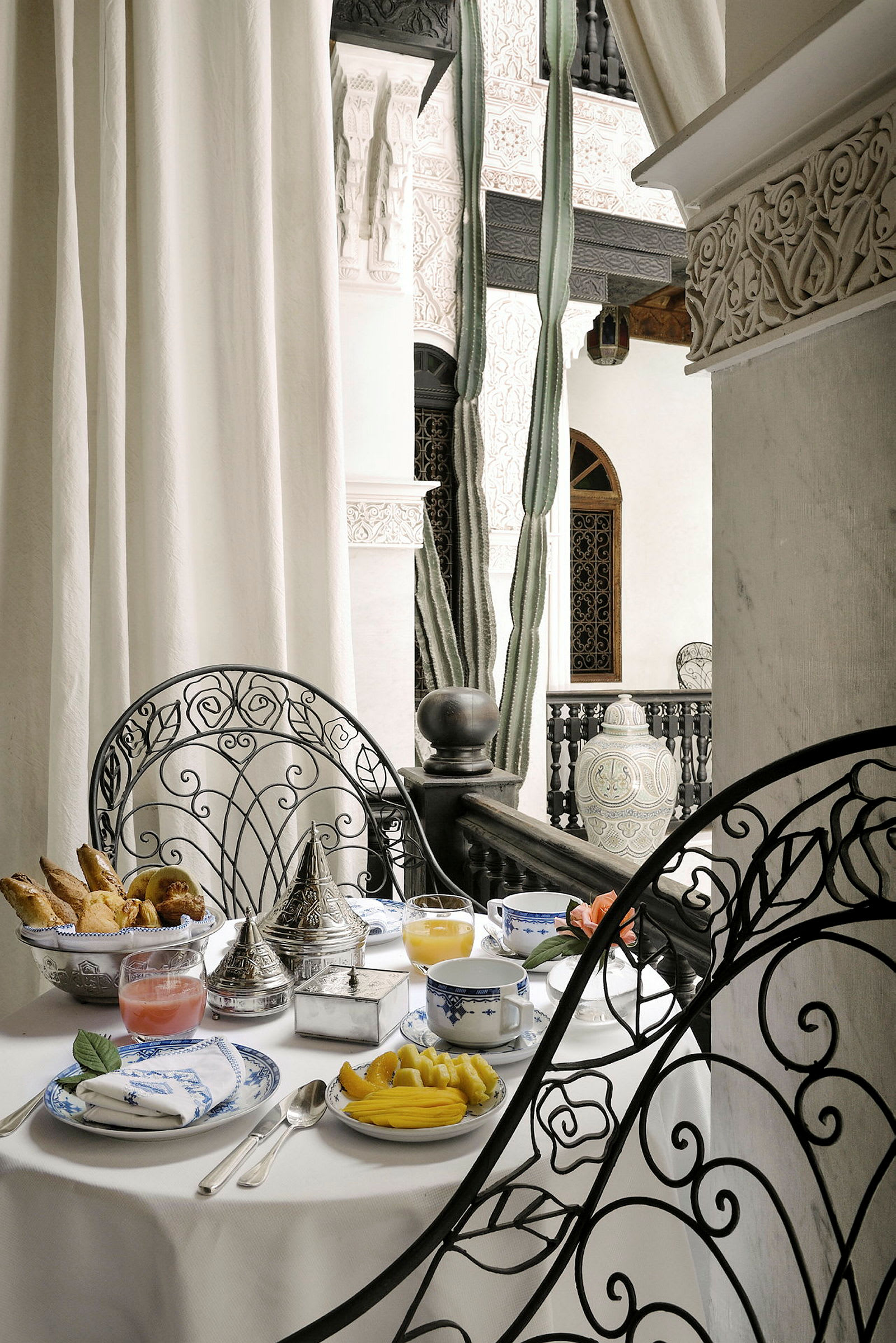 Et bord dækket til morgenmad med porcelæn, drikkevarer og mad, stående på en terrasse med dekoreret metalstol og udsmykninger i orientalsk stil. (AI-genereret tekst)