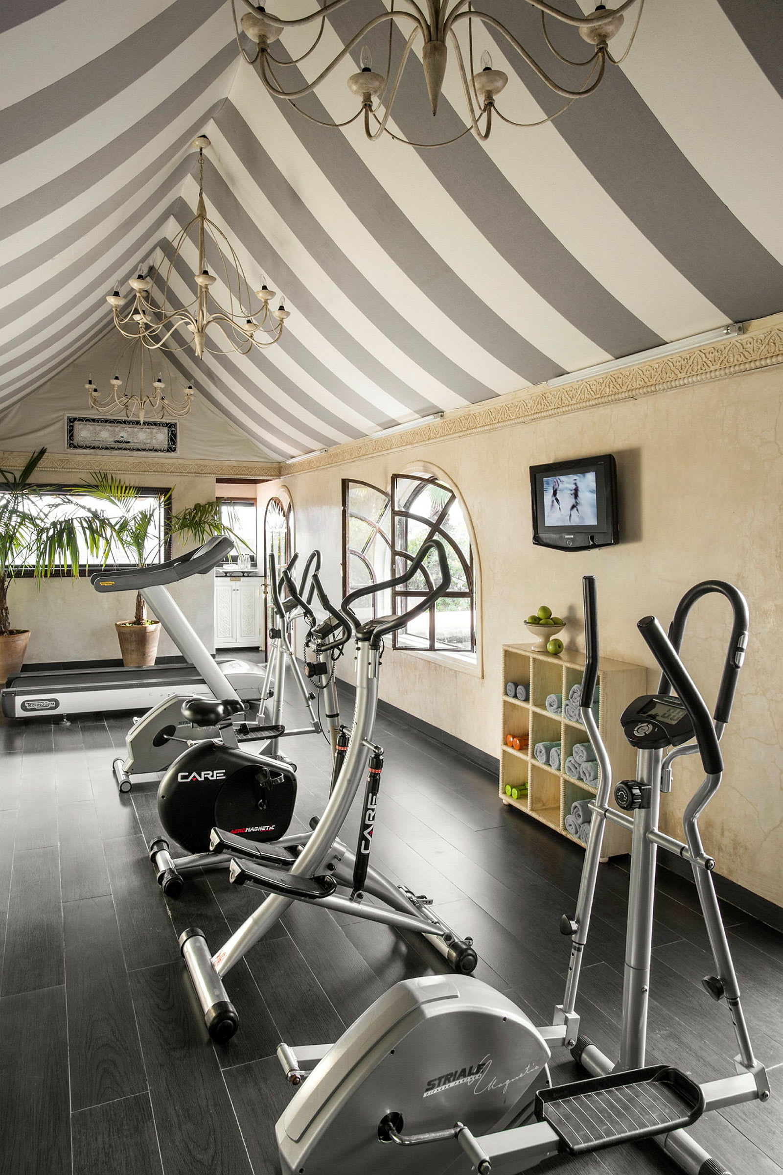 Et fitnessrum med træningsudstyr, herunder motionscykler og løbebånd, i et rum med stribet loft og store vinduer. (AI-genereret tekst)