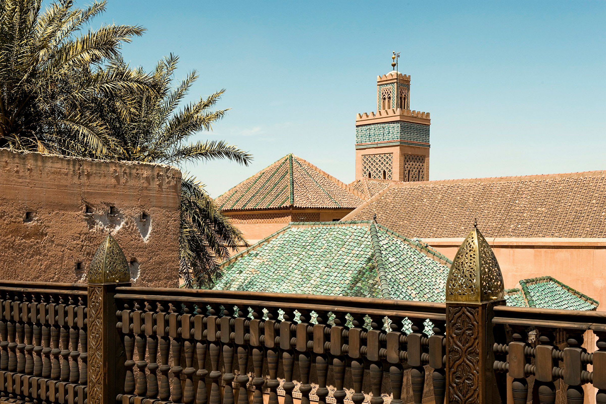 En udsigt over en terrasse med detaljeret rækværk, palmer og en bygning med grønt tag og minaret i baggrunden. (AI-genereret tekst)