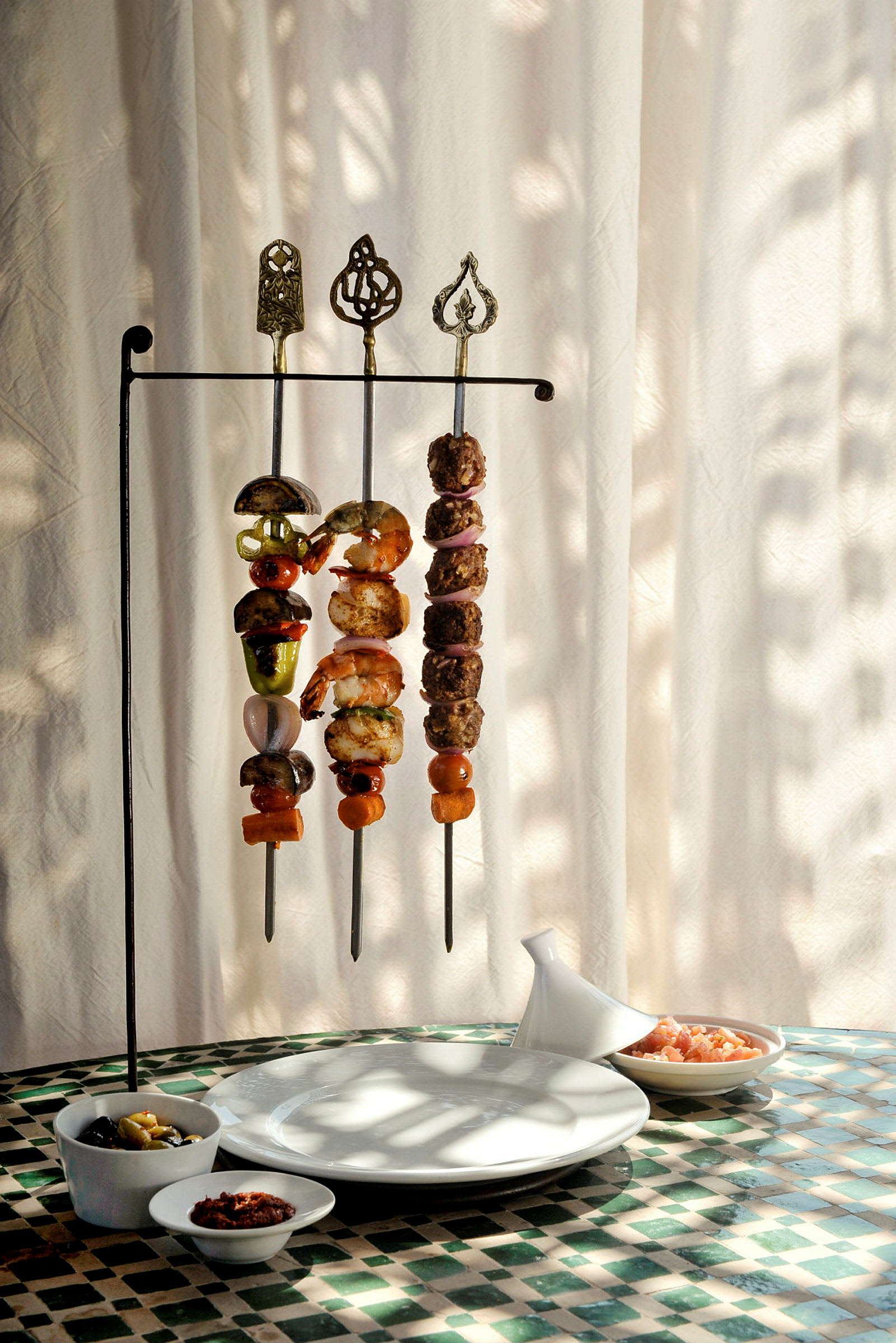 Tre metalskewer med forskellige grillede ingredienser hænger på et metalstativ. Foran stativet står tallerkener og små skåle med forskelligt tilbehør på et bord. (AI-genereret tekst)