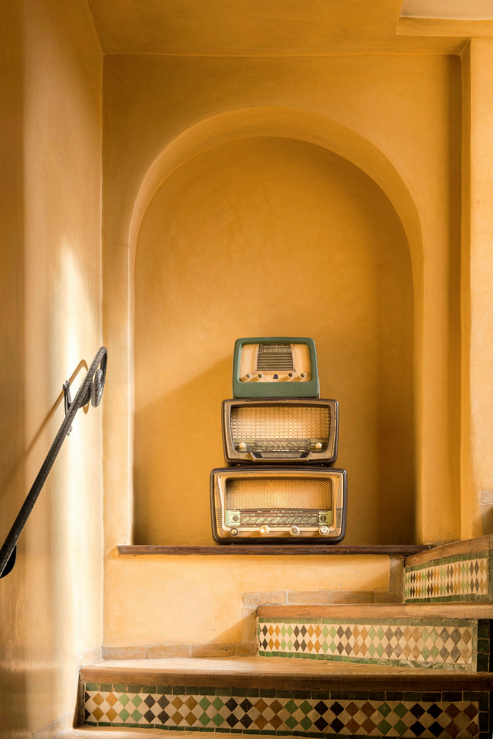 Tre retro-radioapparater er stablet sammen i en indbygget opbevaringsniche ved en trappe med farverige fliser. (AI-genereret tekst)
