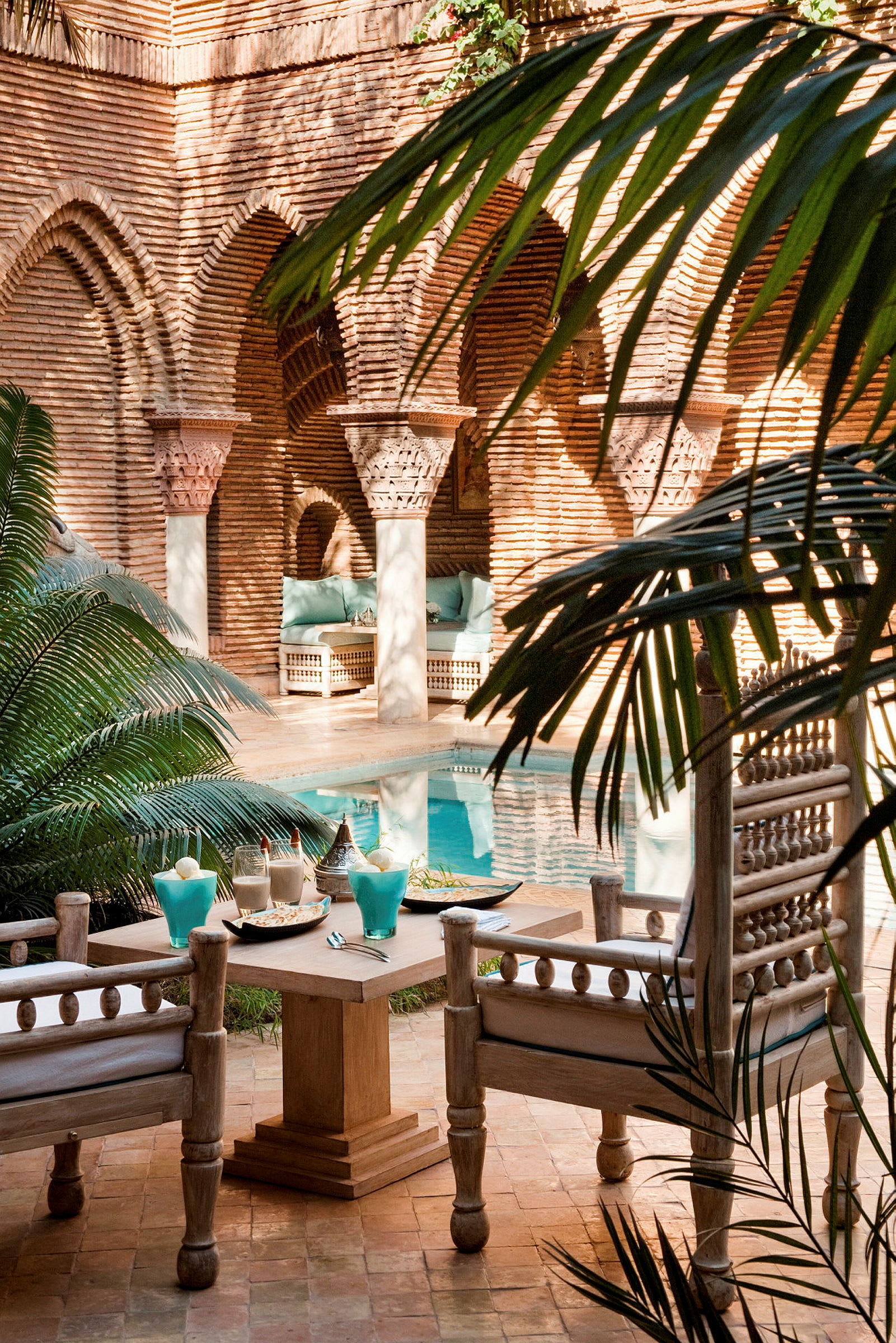 Et siddeområde med bord og stole ved en pool omgivet af planter og arkitektur med buer. (AI-genereret tekst)