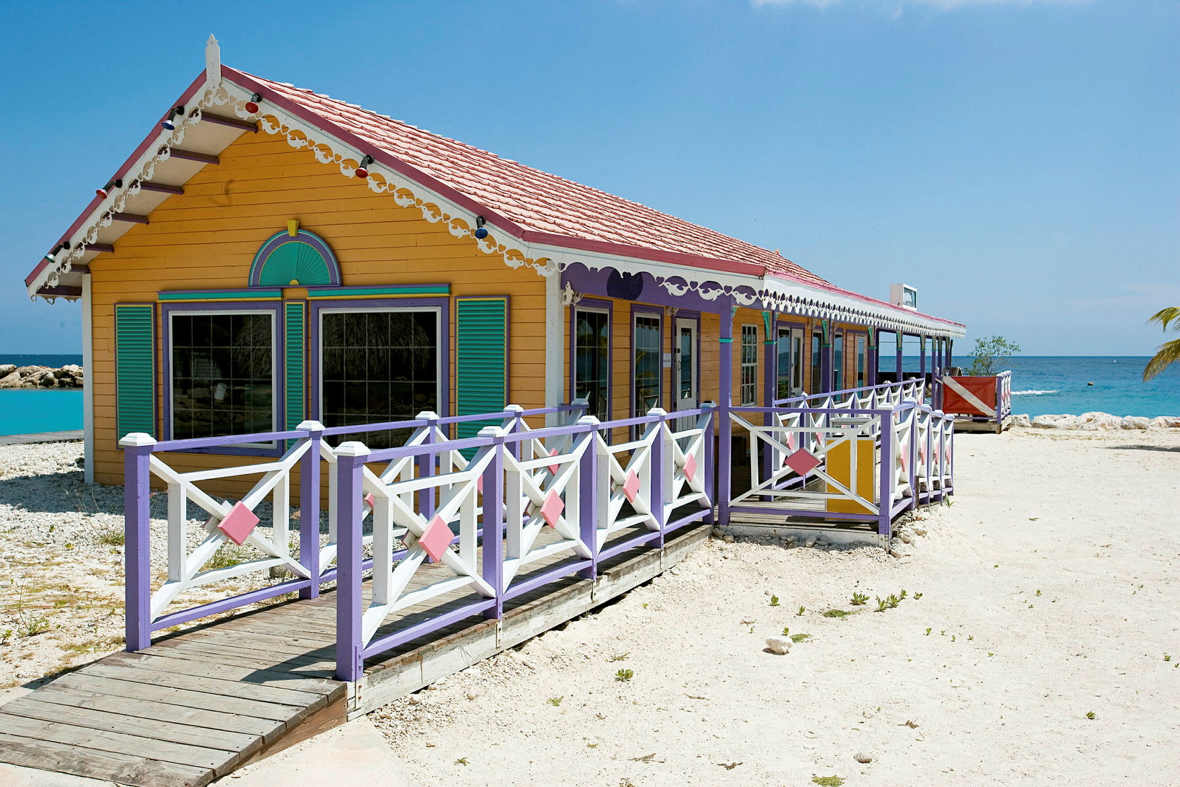 Et farverigt hus med dekorationer og veranda, placeret ved en strand med klart blåt vand. (AI-genereret tekst)