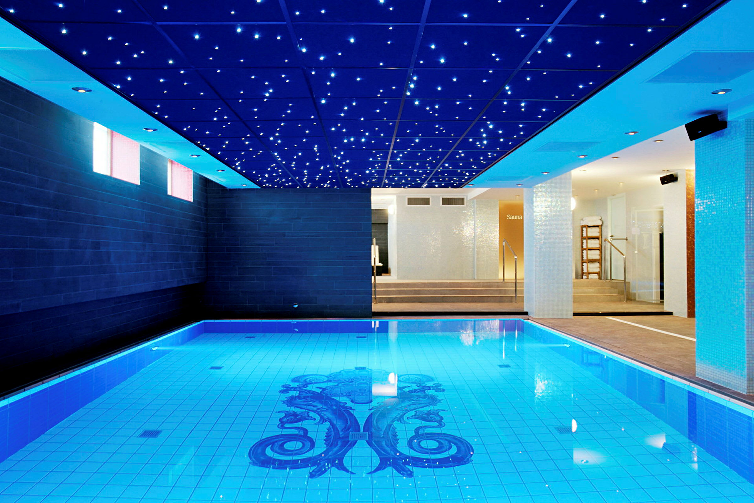 En indendørs swimmingpool med blå flisebelagte vægge og dekorationer på bunden. I loftet er der små lysende lamper, der ligner en stjernehimmel. I baggrunden ses indgangen til en sauna. (AI-genereret tekst)