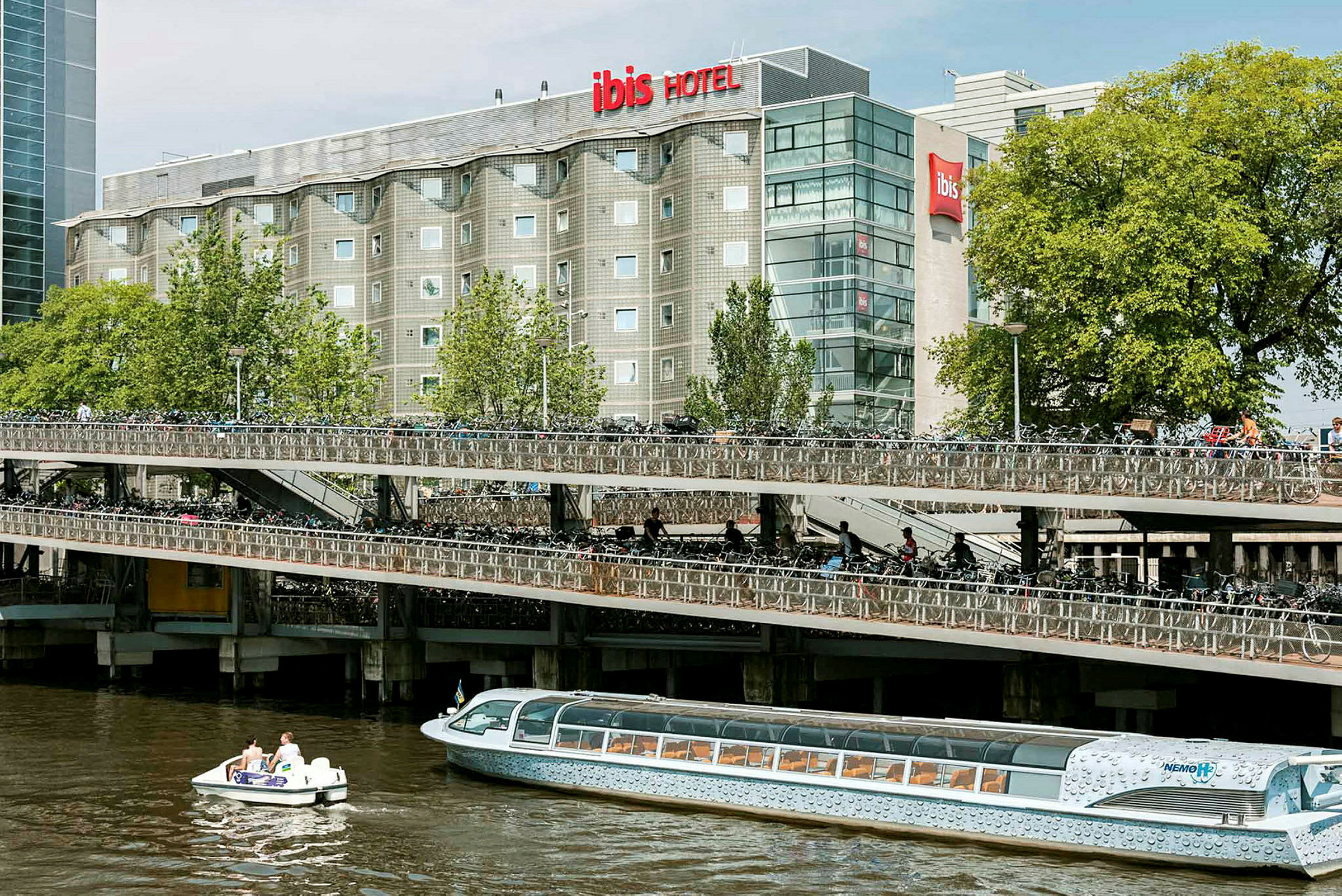 Billede af en treetagers cykelparkering ved siden af en flod, med et hotel i baggrunden og både på vandet. (AI-genereret tekst)