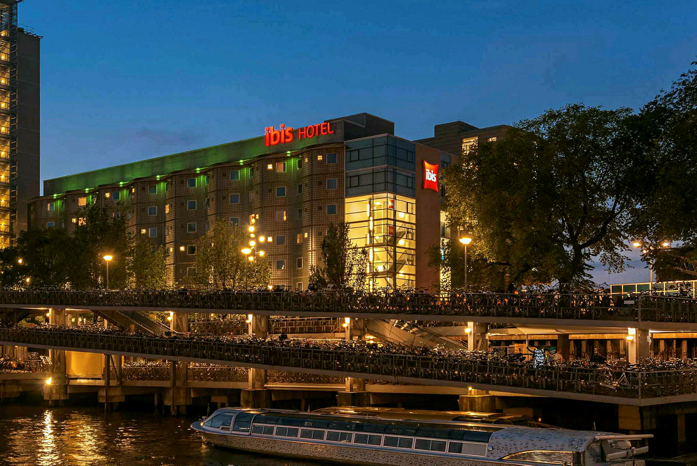 Hotelbygning om aftenen med neonskilt i rødt og grønt, vand og husbåde i forgrunden. (AI-genereret tekst)