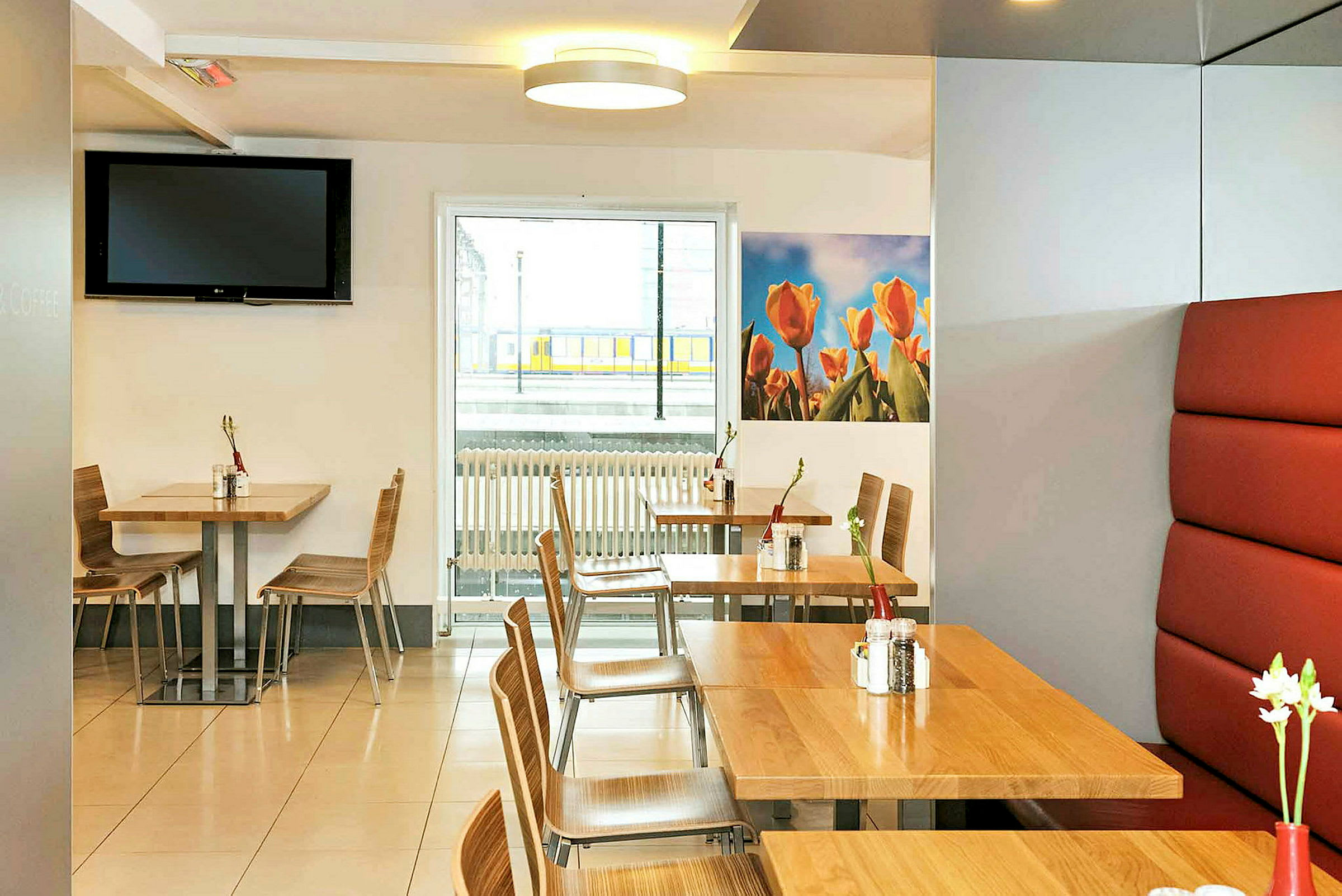 En lys café med borde og stole af træ, dekoreret med små vaser med blomster. På væggen er et billede med tulipaner, og vinduet har udsigt til en togplatform. (AI-genereret tekst)