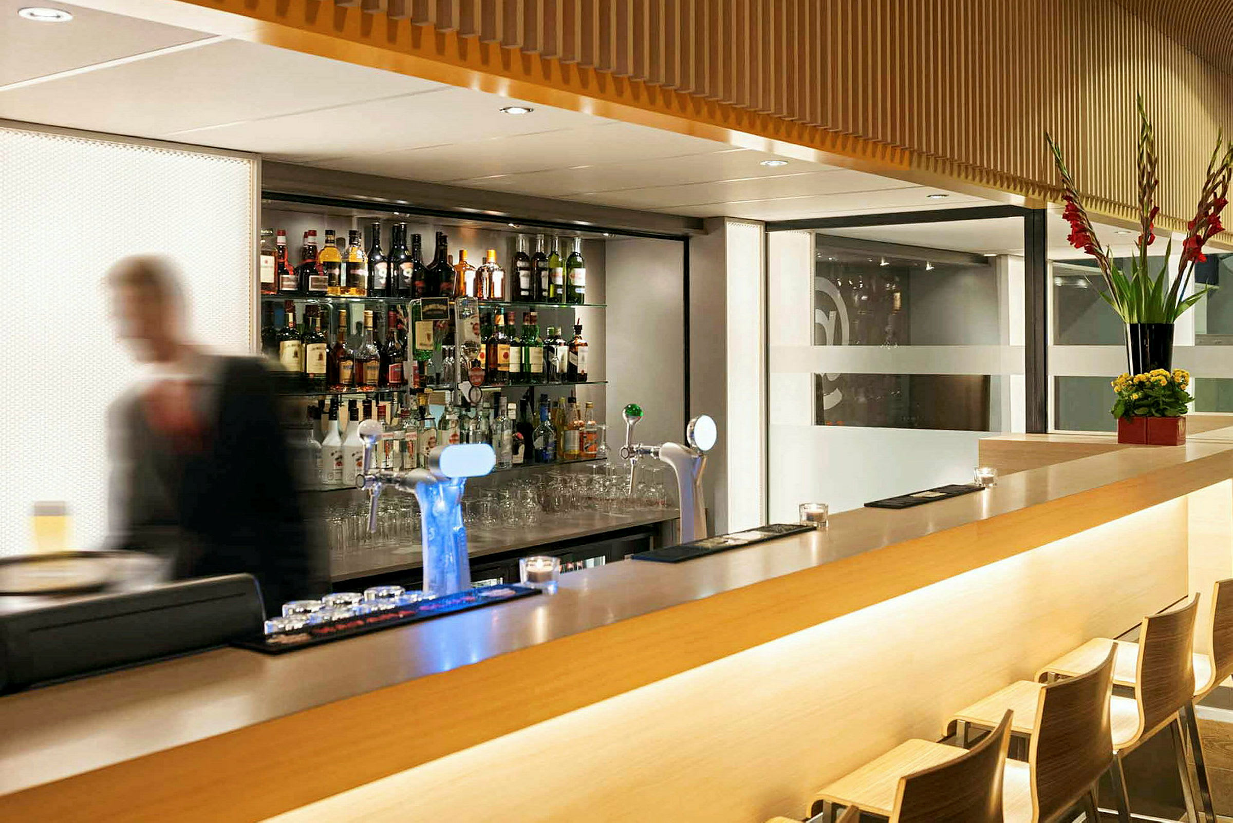 En moderne bar med et udvalg af flasker på hylder bagved samt en sløret person i forgrunden. (AI-genereret tekst)
