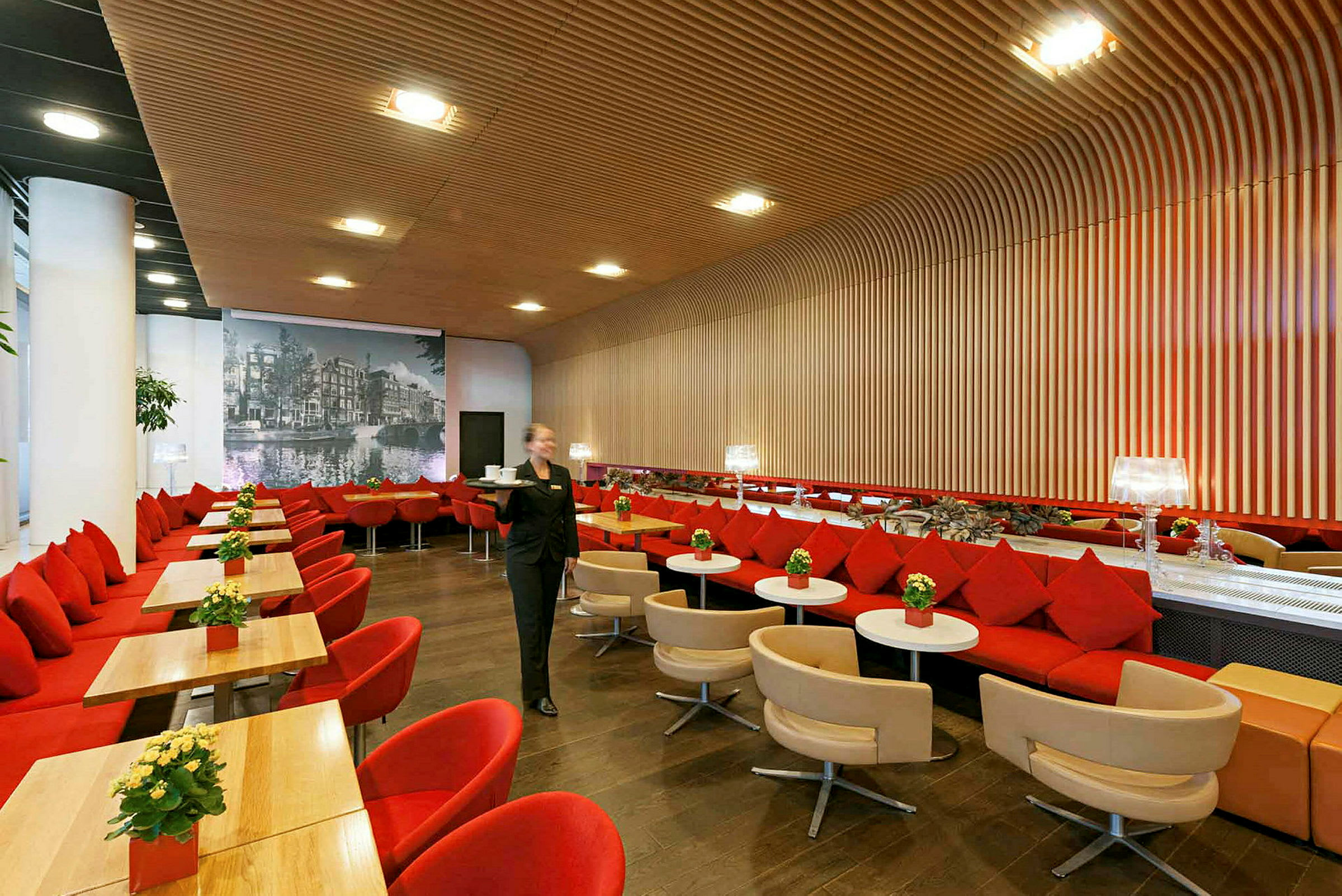 En moderne restaurant med røde og beige siddepladser, en stor væg med træpaneler og et billede af byen i baggrunden. Bord og stole er arrangeret i rækker. (AI-genereret tekst)