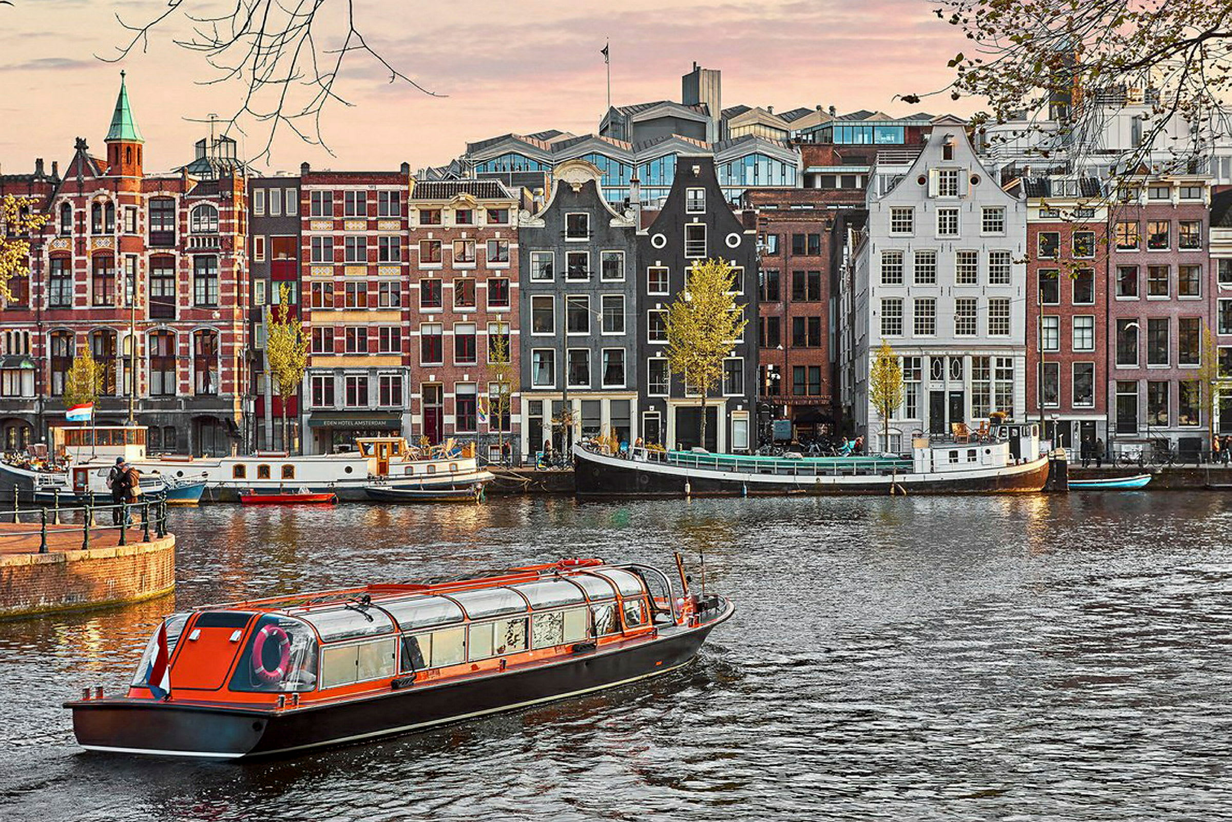 Flere bygninger med traditionelle facader ved en kanal, hvor en sightseeingbåd passerer i forgrunden. (AI-genereret tekst)