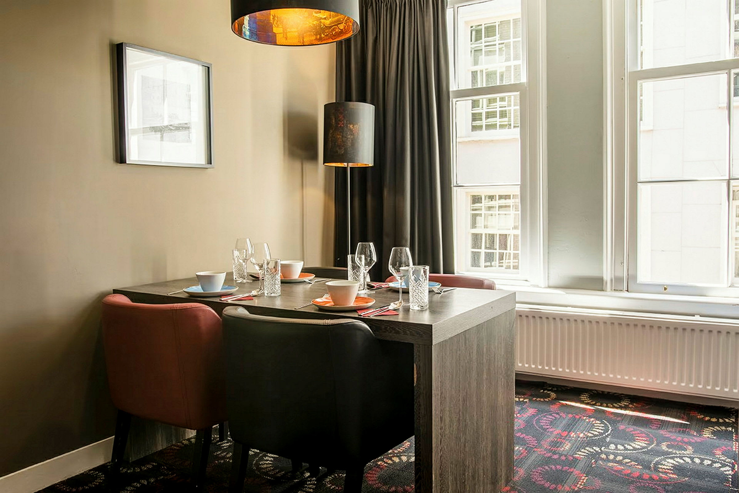 Et spisebord med fire stole. Bordet er dækket med glas, tallerkener og skåle. En orange hængelampe lyser over bordet. Vinduer og gardiner ses i baggrunden. (AI-genereret tekst)