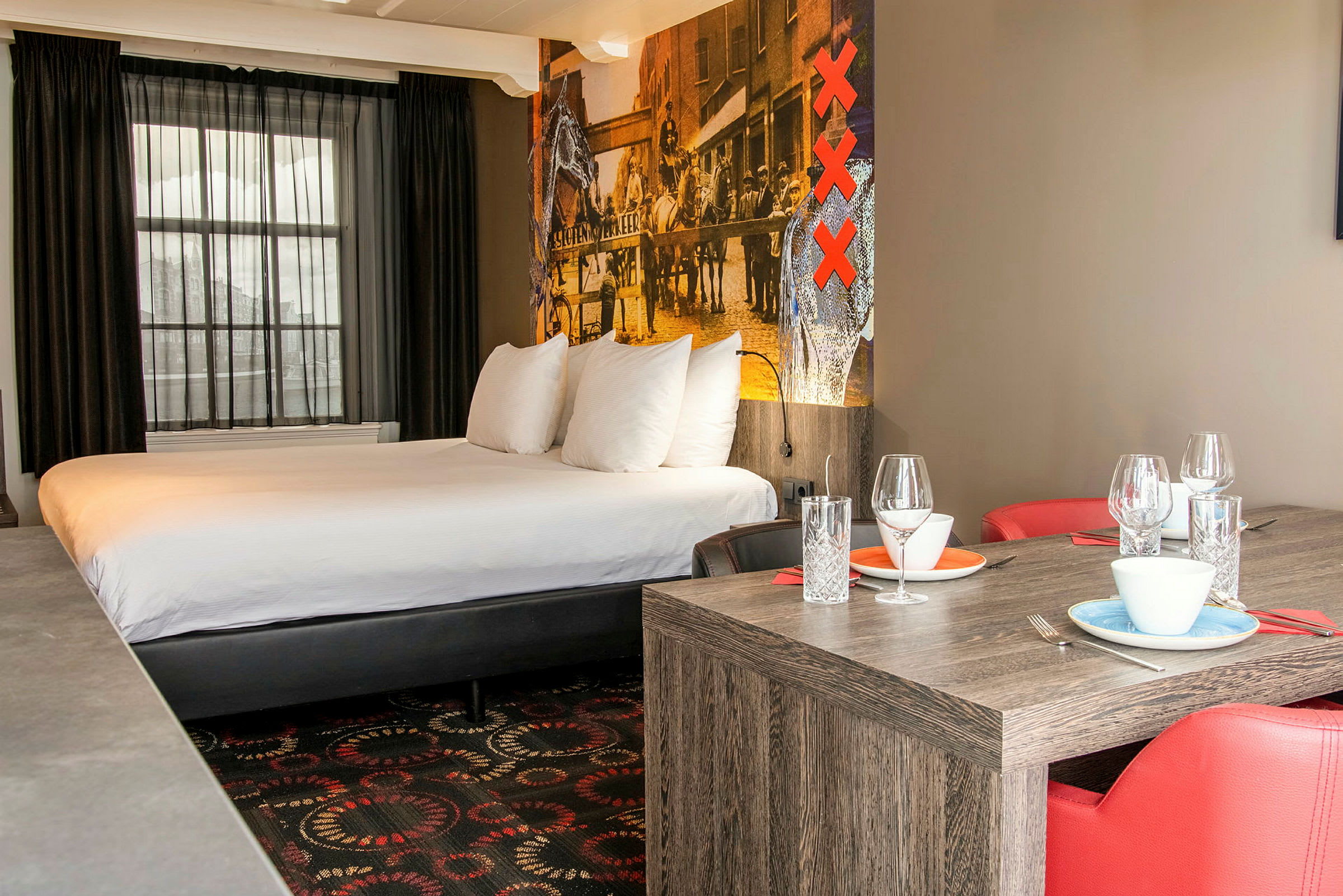 Et hotelværelse med en stor seng, røde stole og et bord med dækket service og glas. (AI-genereret tekst)