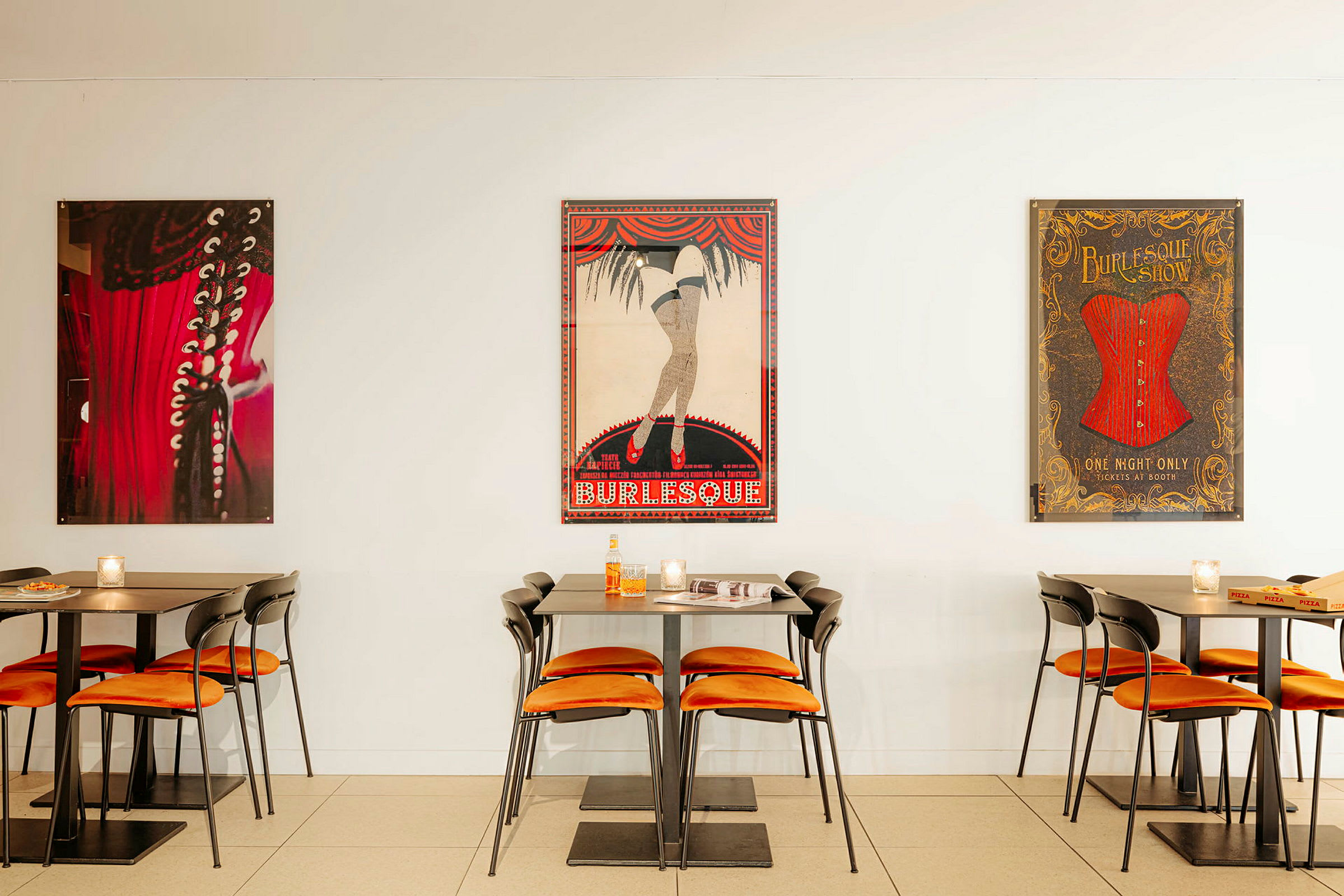 Billede af et cafémiljø med borde og stole samt burlesque-inspirerede plakater på væggen. (AI-genereret tekst)