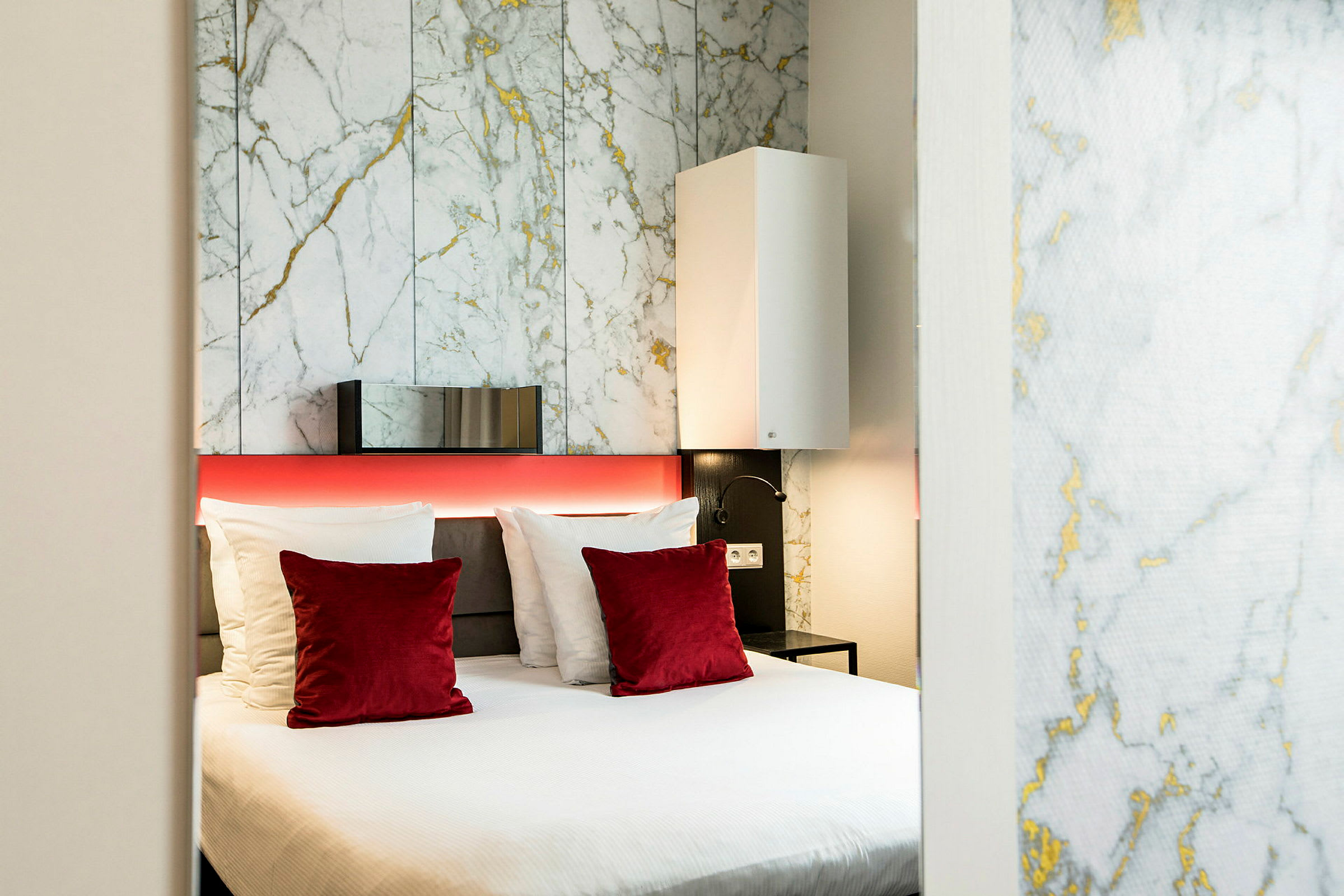 Et moderne hotelværelse med en dobbeltseng, hvide og røde puder samt en bagvæg med marmormønster. (AI-genereret tekst)