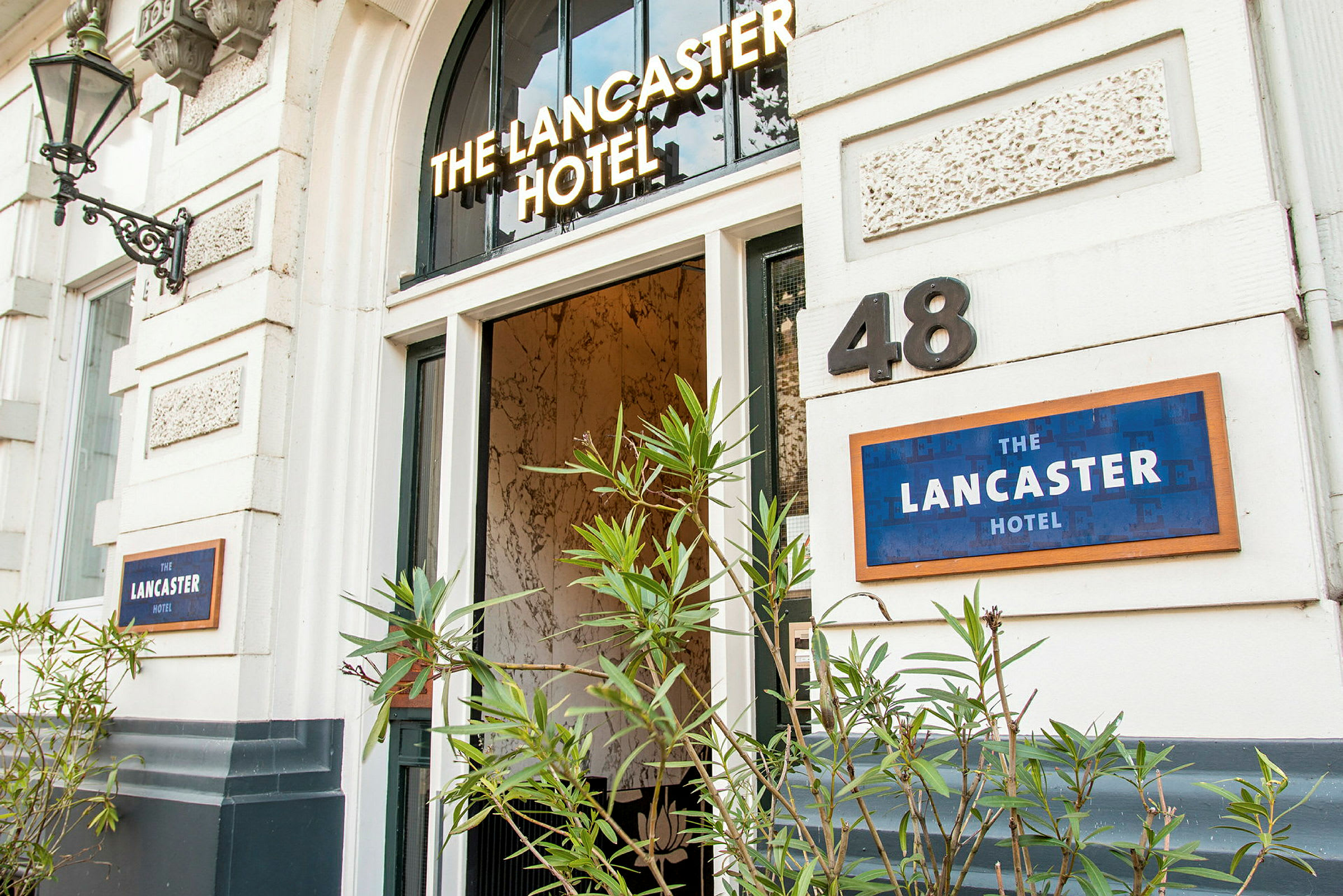Indgang til et hotel med navnet 'The Lancaster Hotel', omgivet af grønne planter. (AI-genereret tekst)