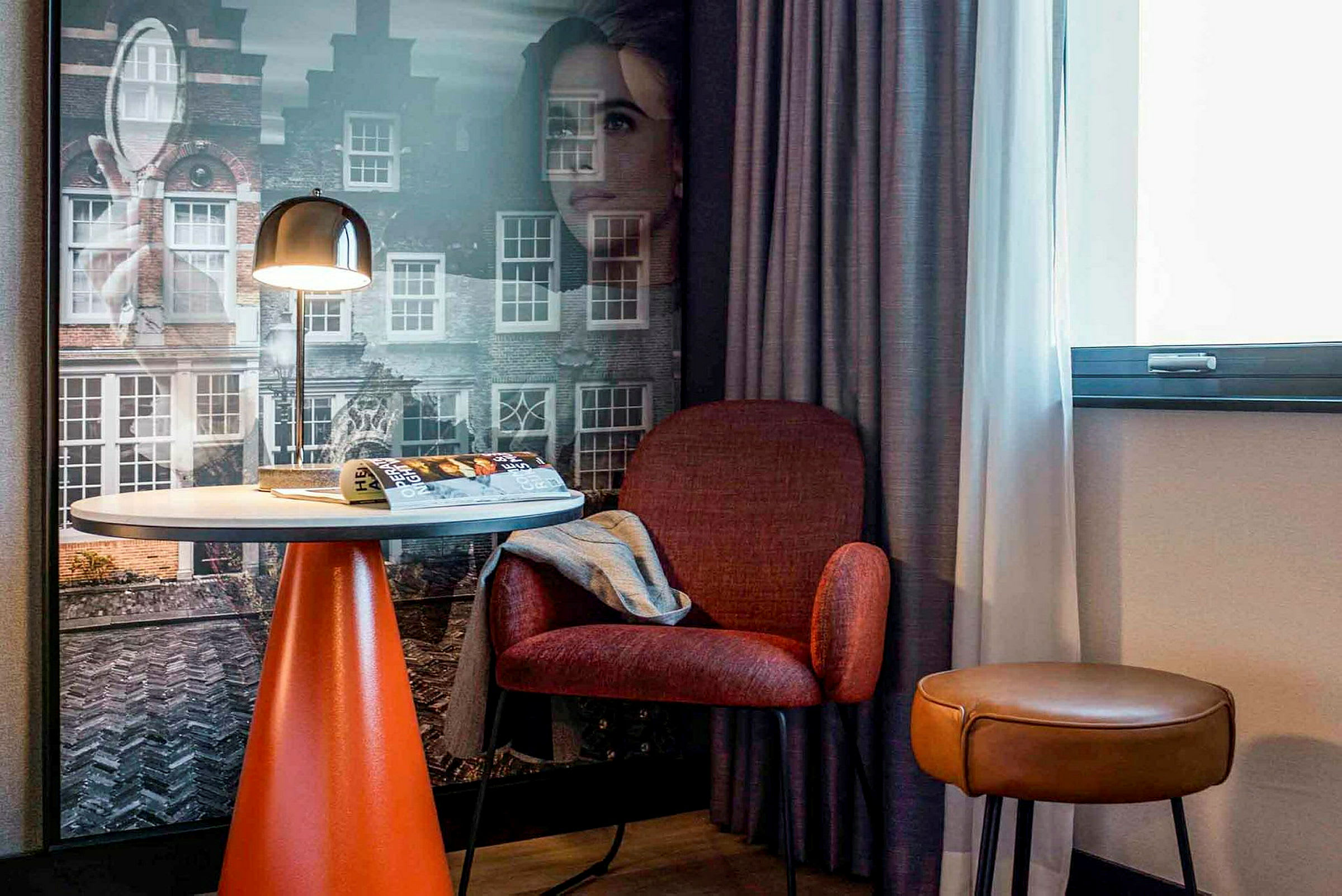 Et hotelværelse med et bord, en rød stol og en brun skammel foran et vindue og en væg med bymotiver. (AI-genereret tekst)