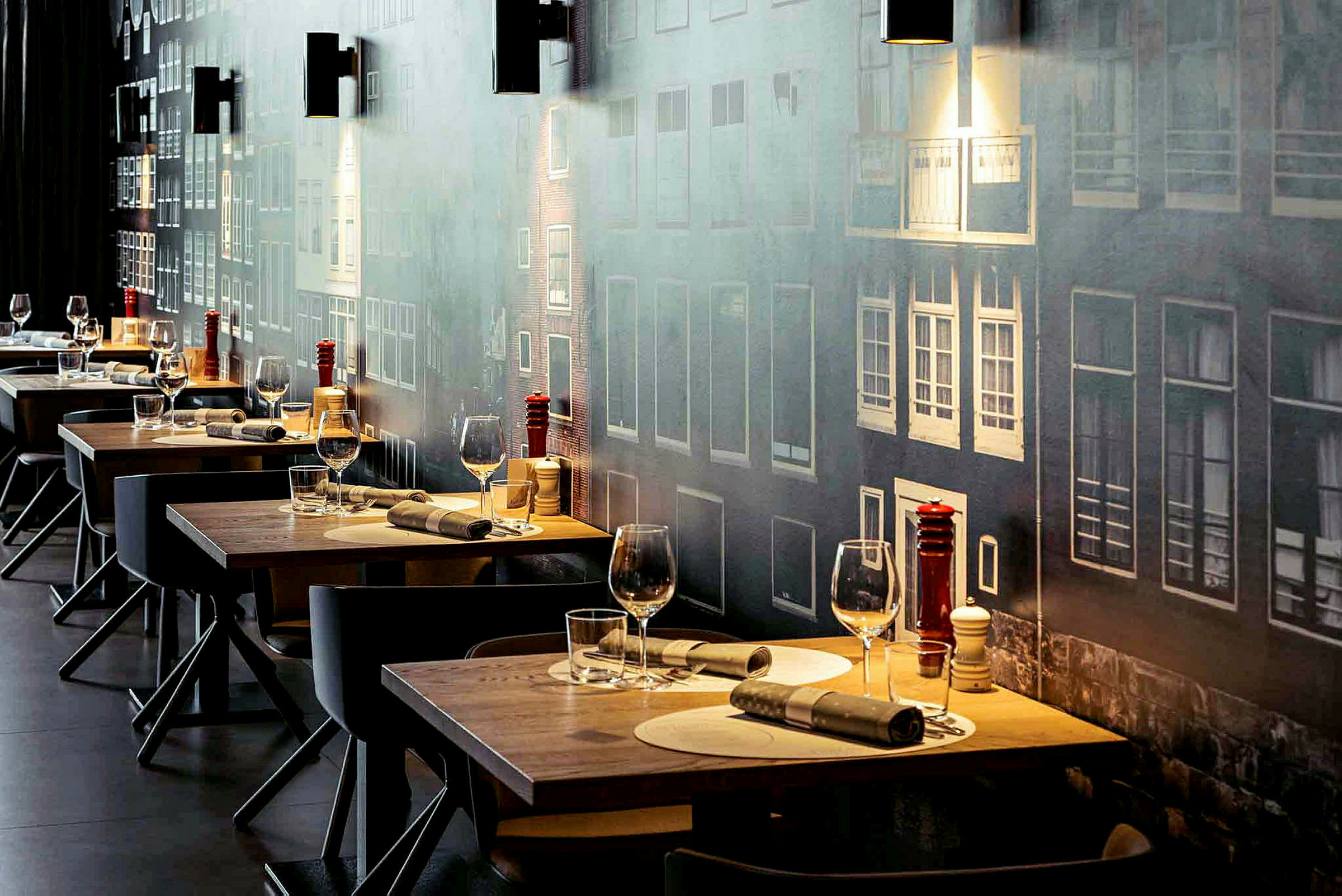 Flere dækkede restaurantborde med glas og bestik, mod en væg med facademotiv. (AI-genereret tekst)