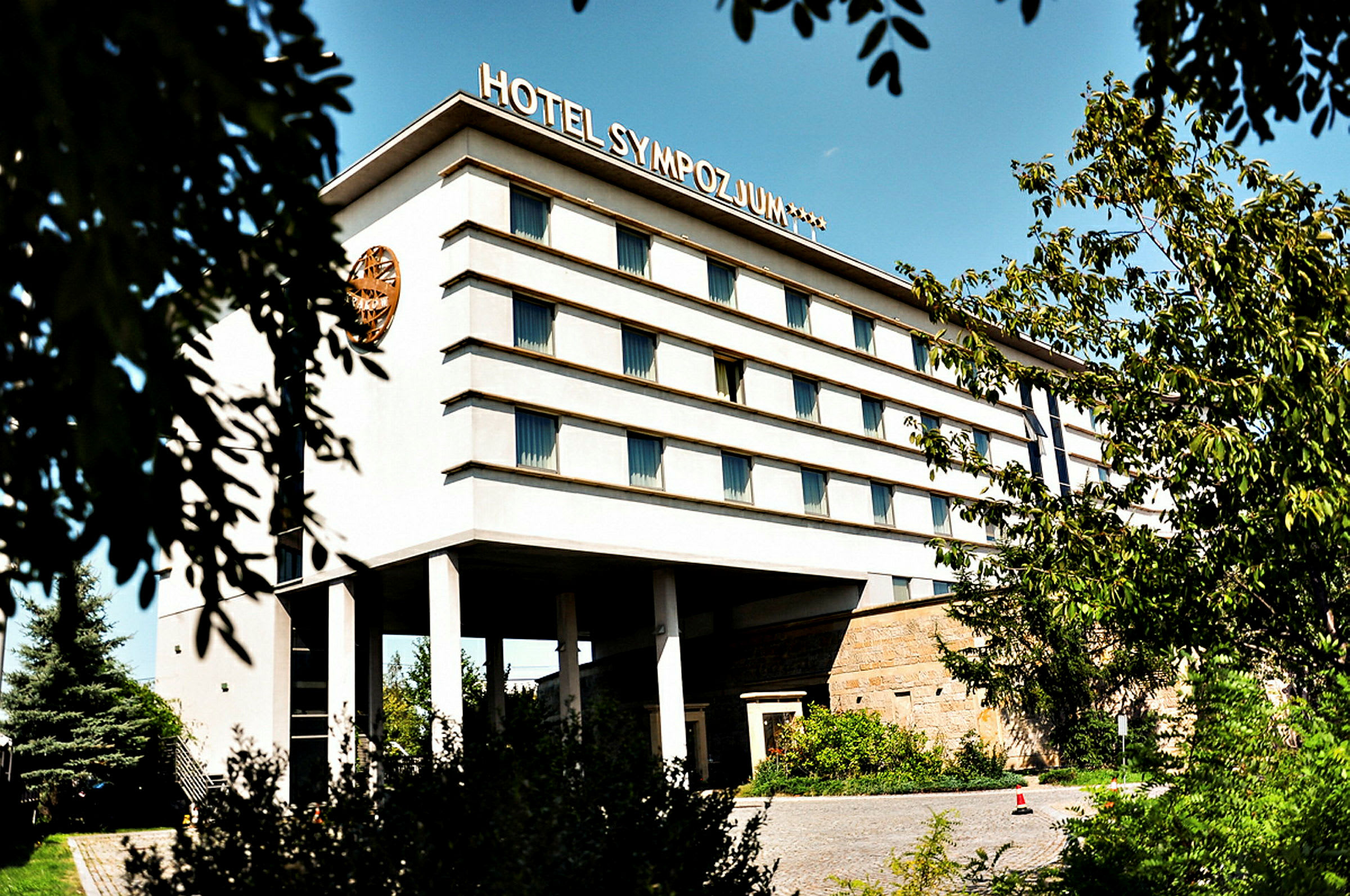 Bygning med flere etager, mærket med teksten 'Hotel Sympozjum', omgivet af træer og vegetation. (AI-genereret tekst)