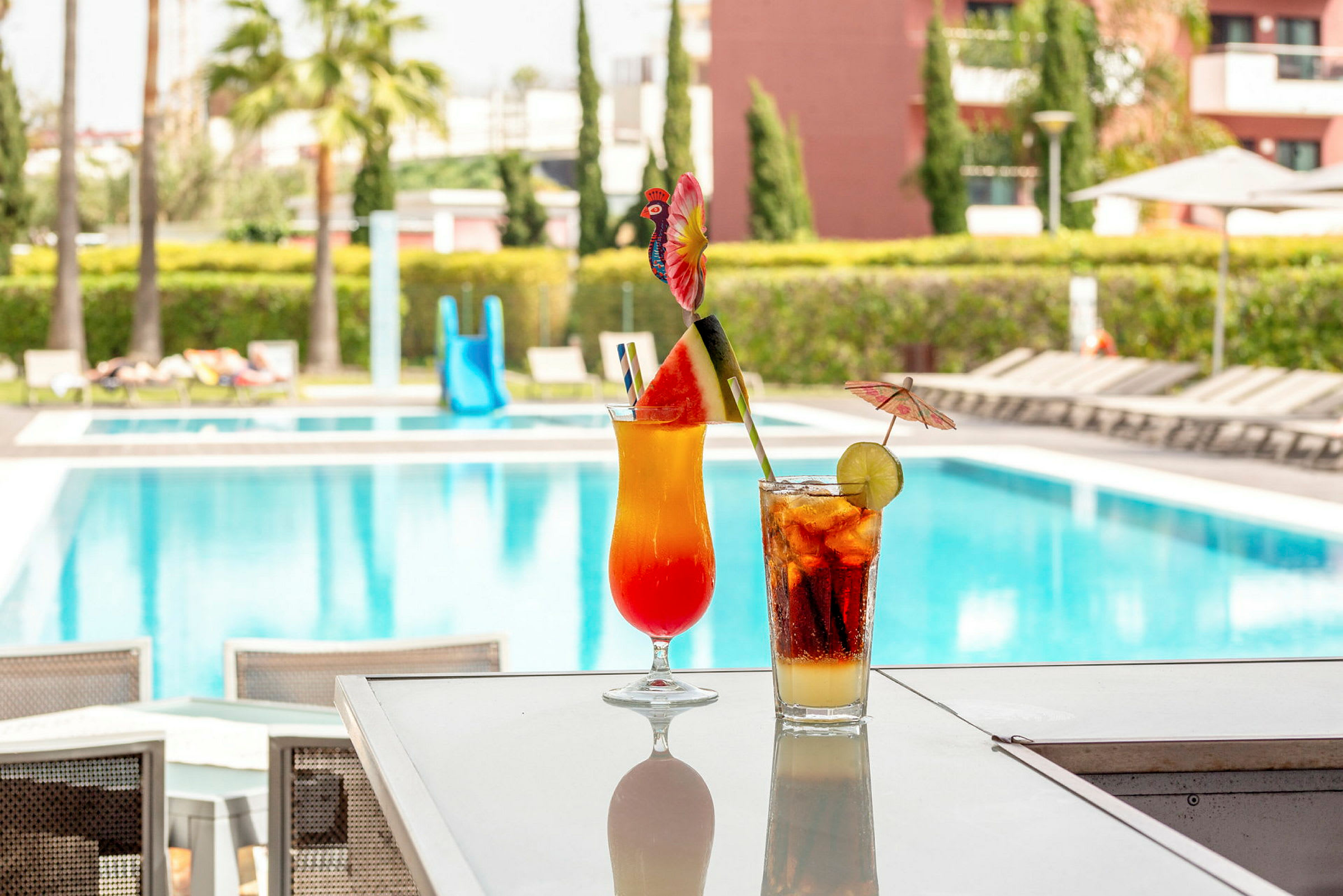 To farverige cocktails på et bord med en swimmingpool i baggrunden. (AI-genereret tekst)
