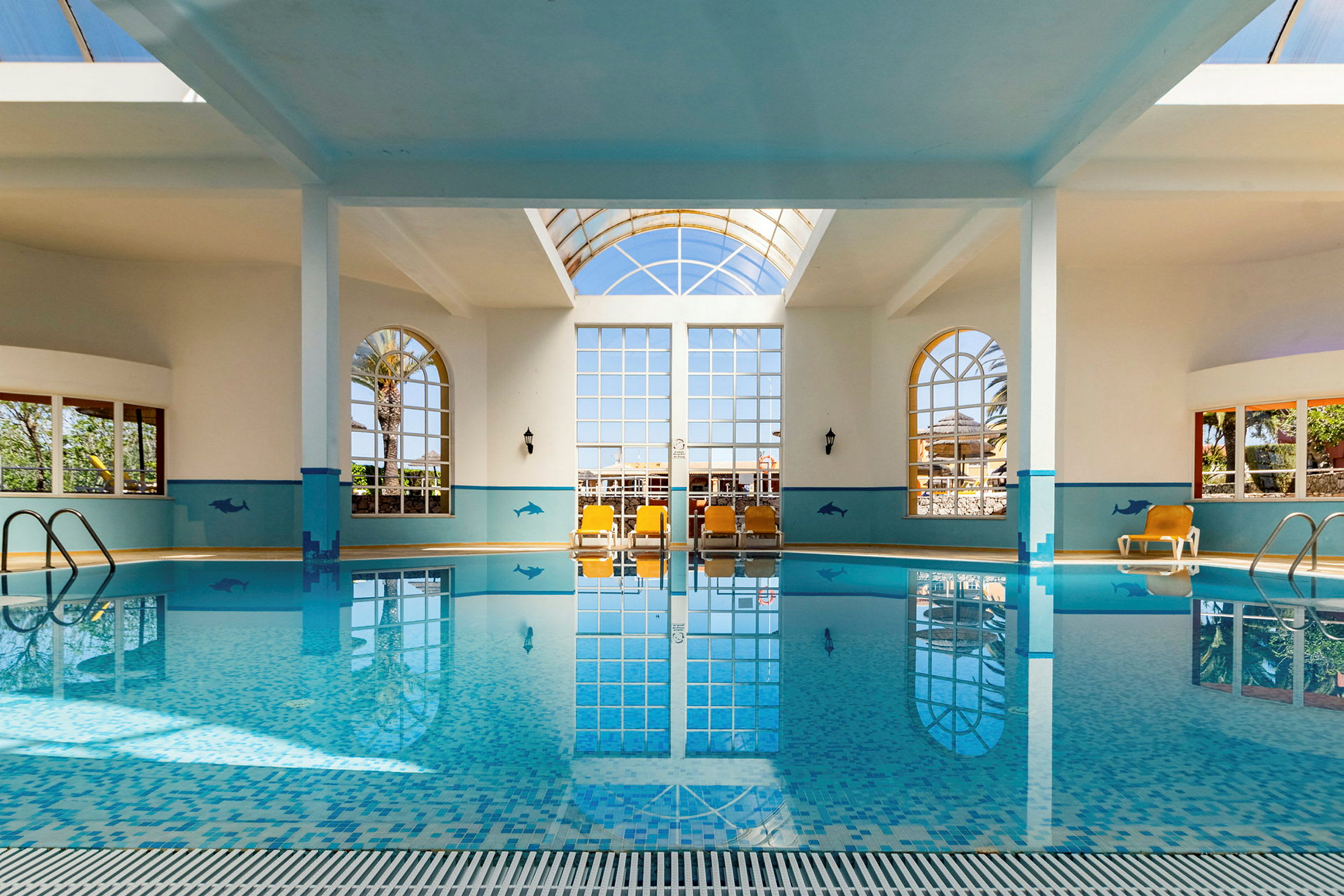 Indendørs pool med store vinduer, solsenge og dekorative vægmalerier. (AI-genereret tekst)