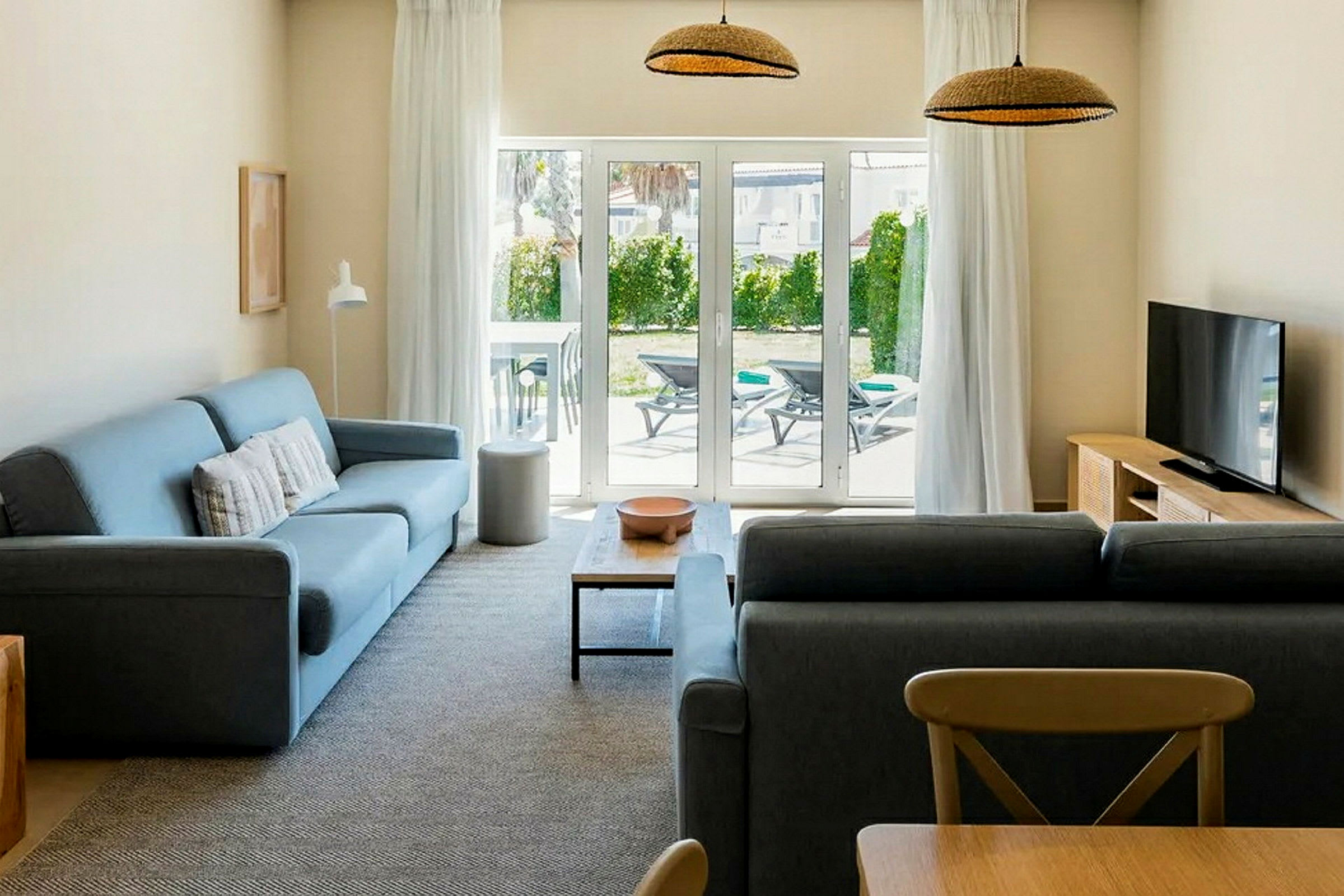 En stue med to sofaer, et sofabord, et fjernsyn og en glasdør, der fører ud til en terrasse med liggestole. (AI-genereret tekst)