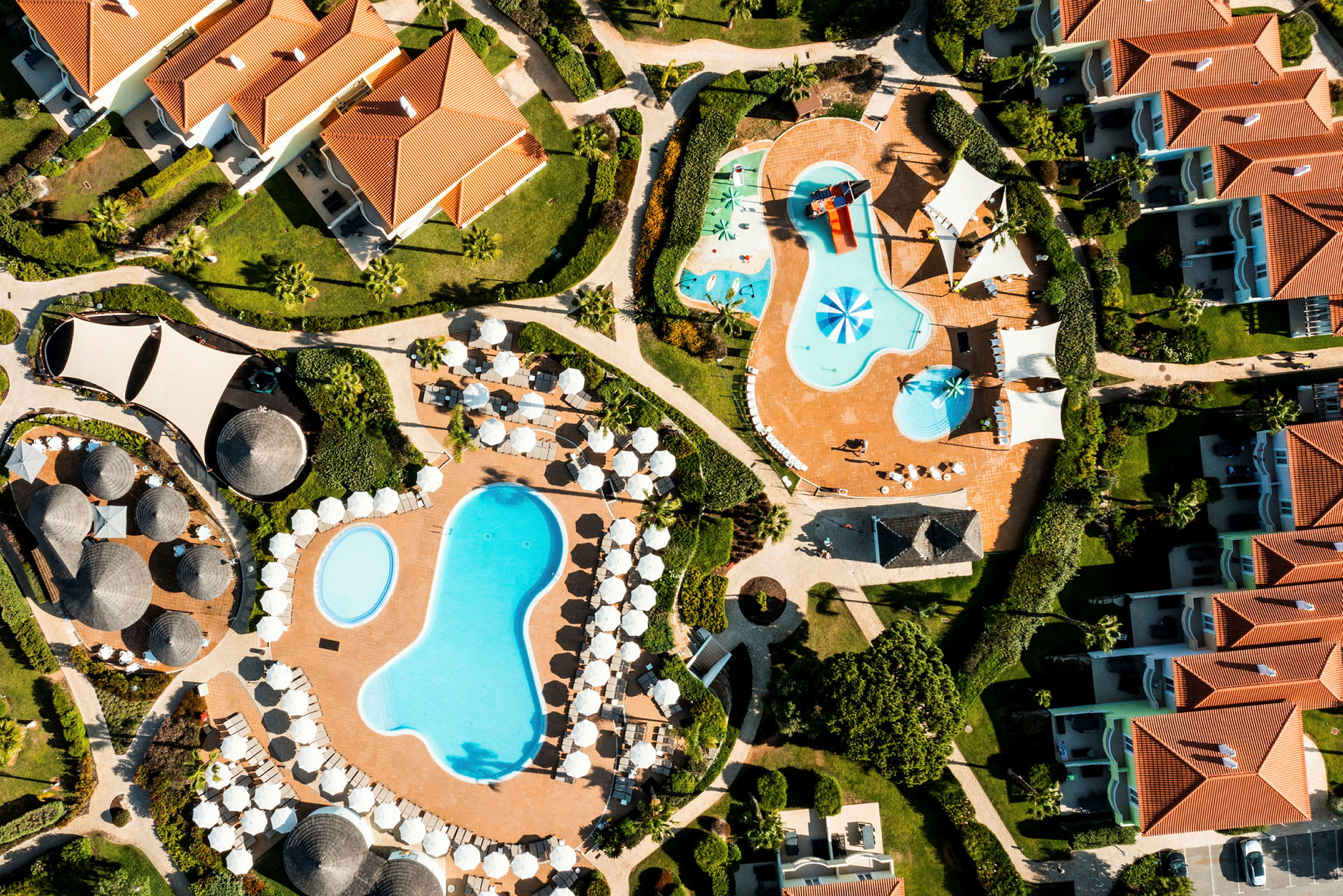 Luftfoto af resortområde med flere pools, liggestole, parasoller og bygninger. (AI-genereret tekst)