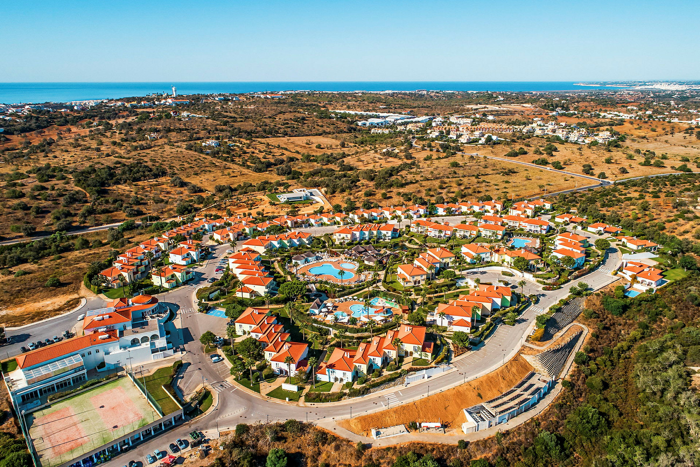 Luftfoto af et feriested med røde tage, omgivet af landskab og havet i baggrunden. (AI-genereret tekst)