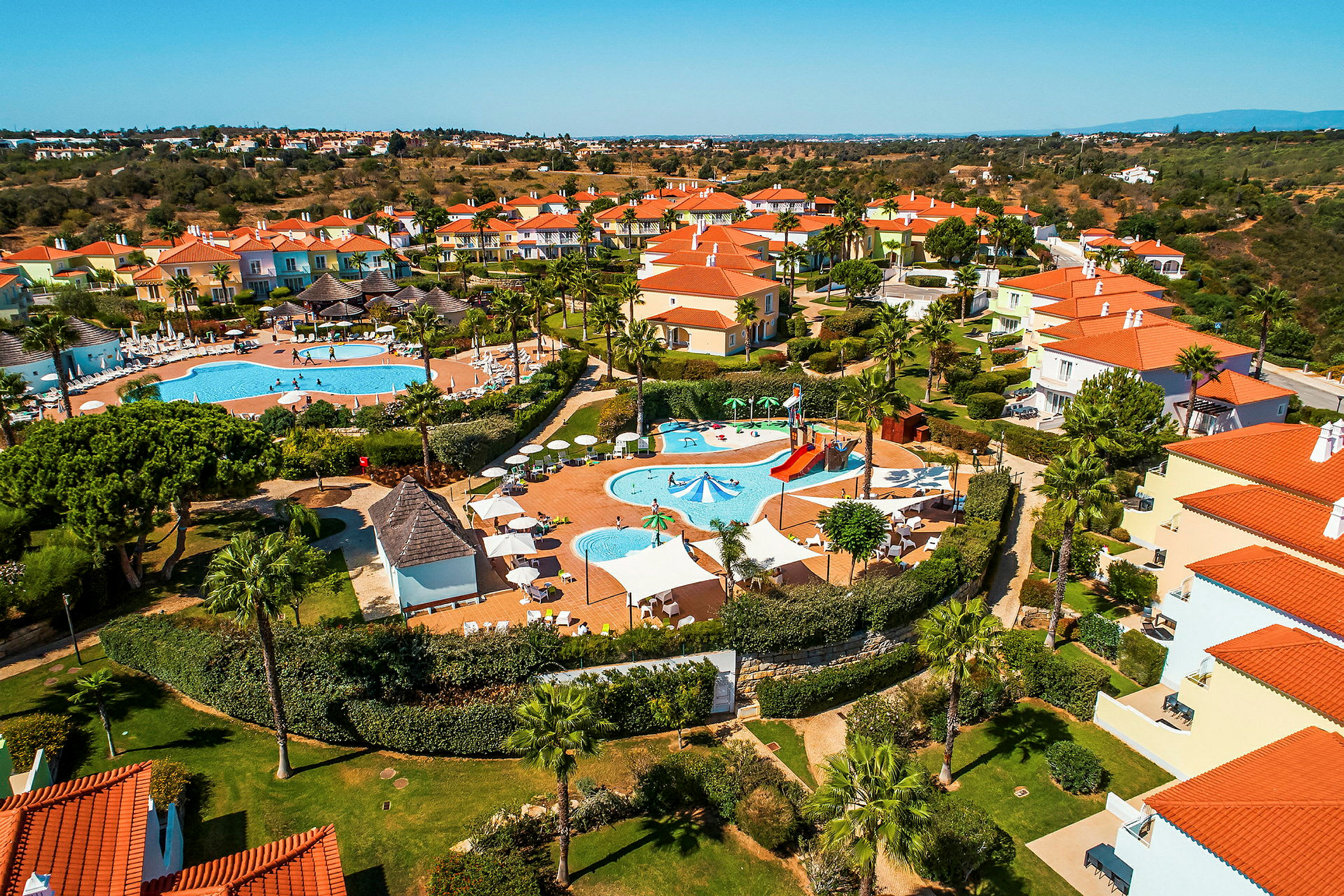 Luftfoto af et feriekompleks med pools og farverige huse. (AI-genereret tekst)