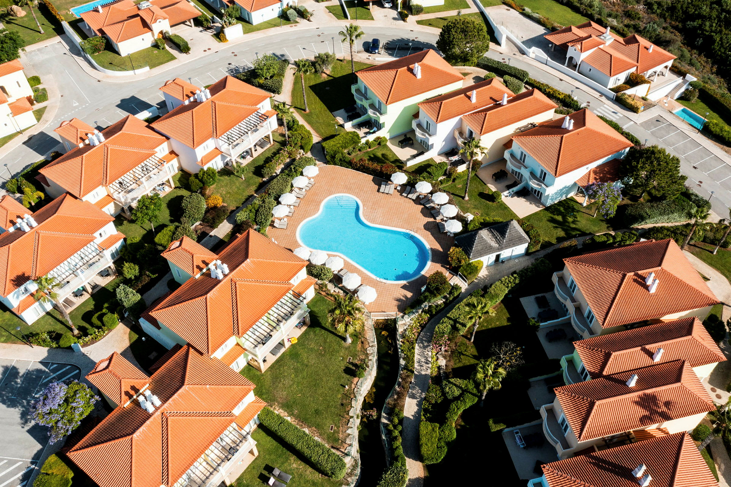 Luftfoto af ferieboliger med røde tage omkring en pool. (AI-genereret tekst)
