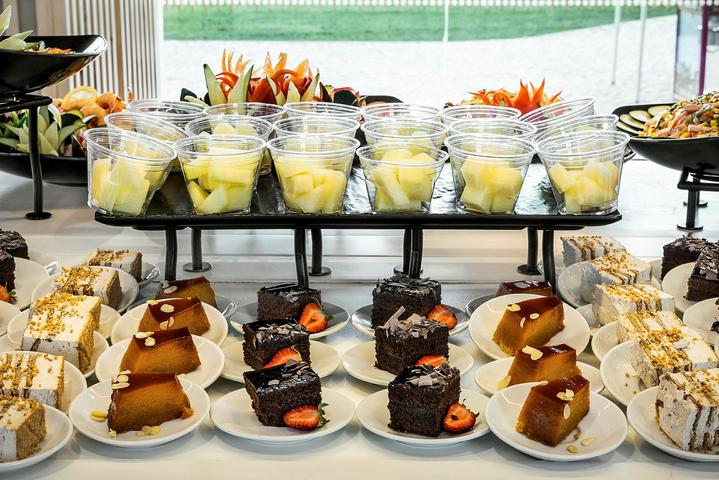 Buffet med desserter og frugtsalat arrangeret på tallerkener og i plastglas. (AI-genereret tekst)