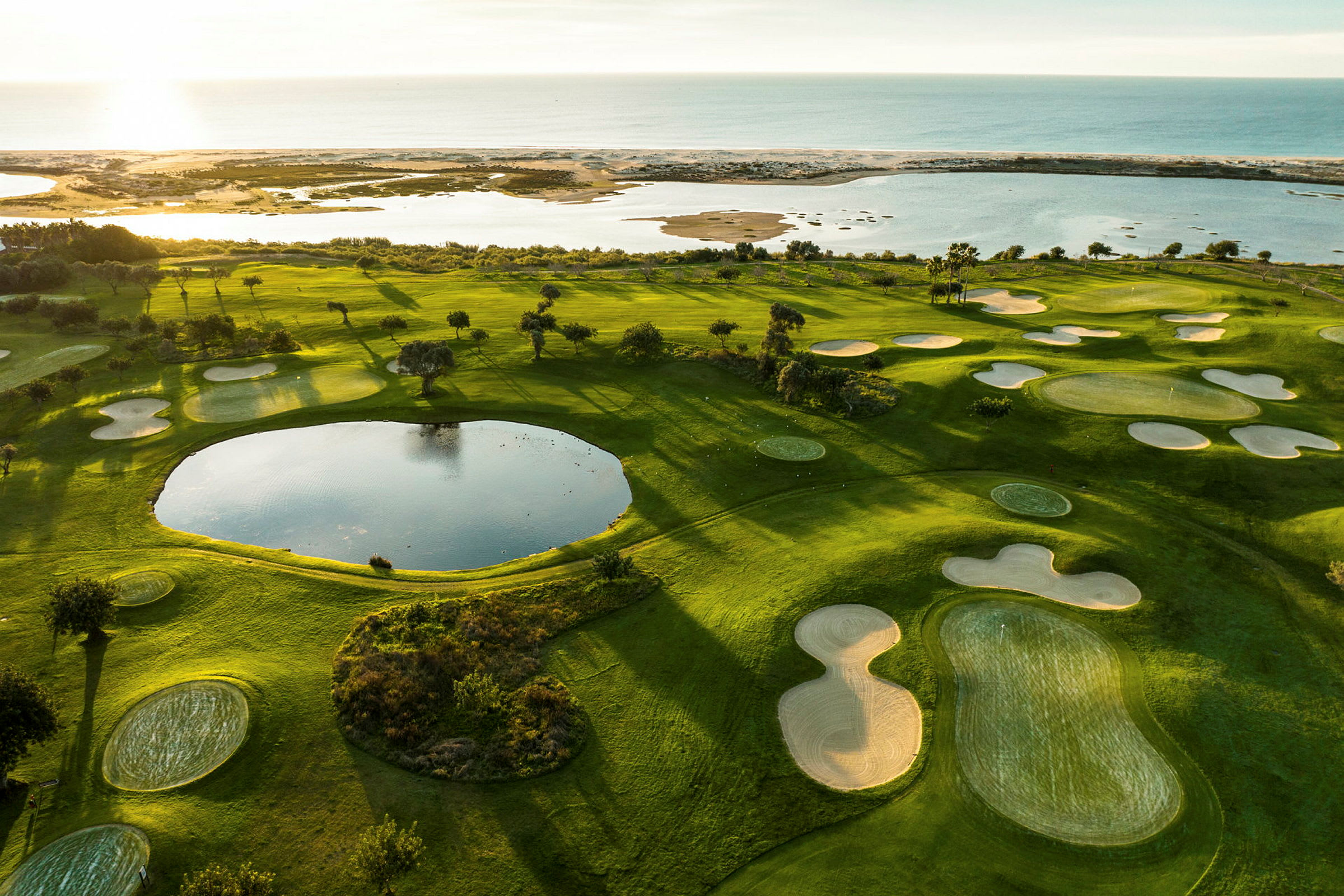 Udsigt over en grøn golfbane med sandbunkere og en dam, tæt på kysten og havet. (AI-genereret tekst)