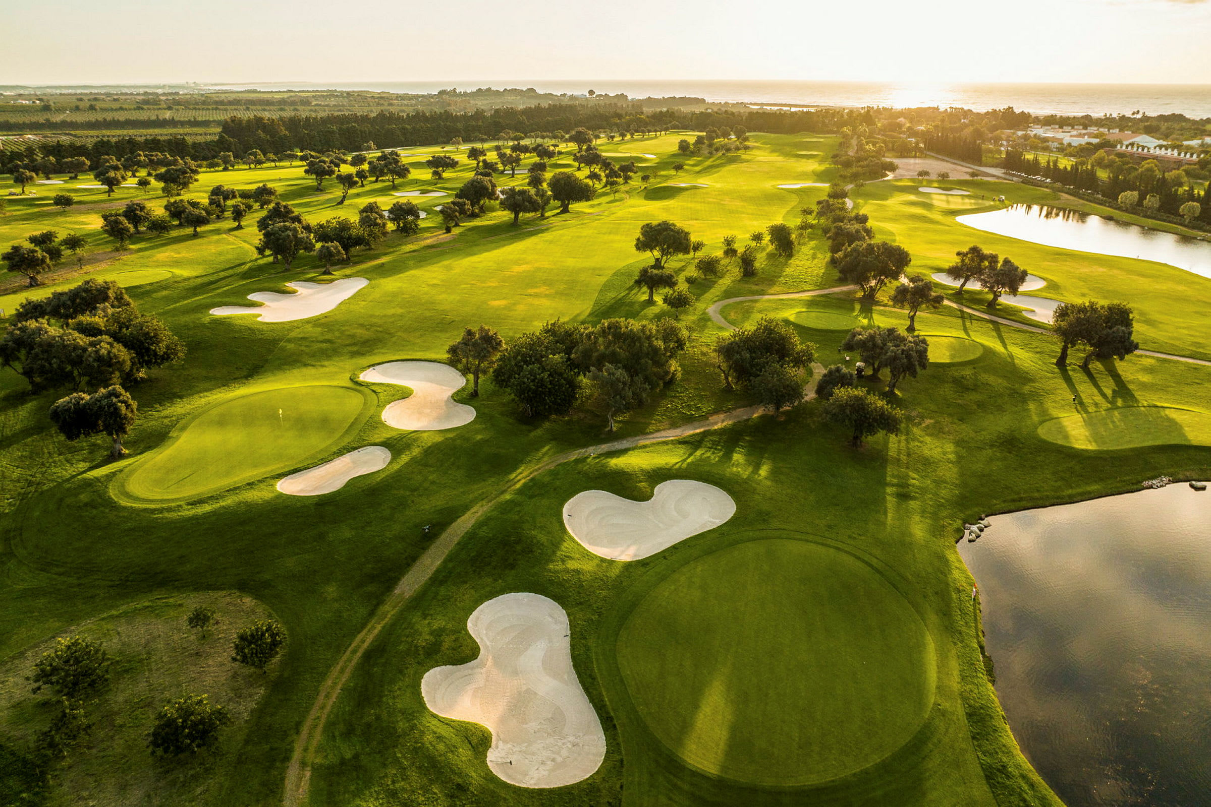 Luftudsigt over en golfbane med grønne fairways, sandbunkers og vandhazarder ved solnedgang. (AI-genereret tekst)