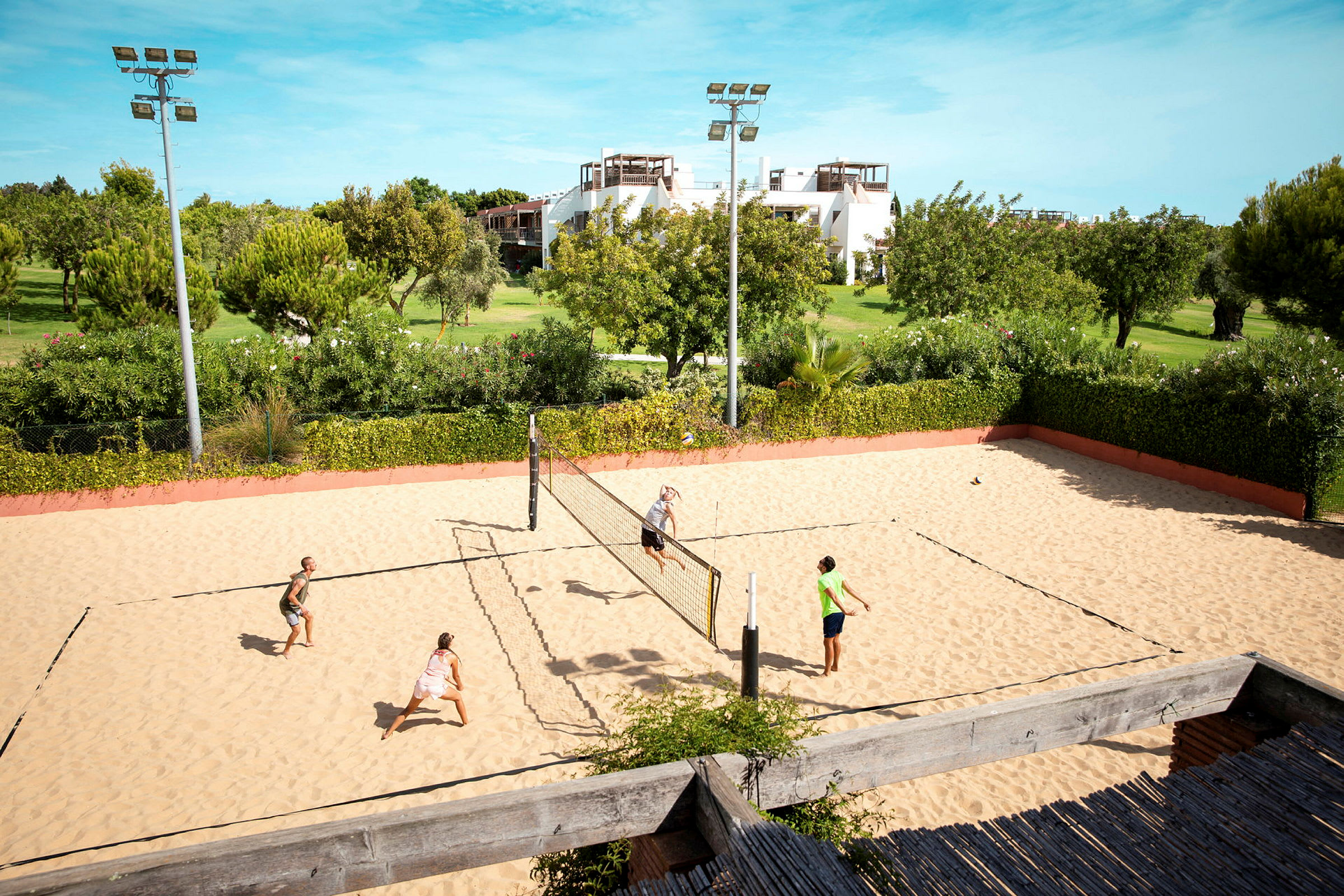 Sandvolleyballbane med fire personer, der spiller, omgivet af grønt landskab og bygninger i baggrunden. (AI-genereret tekst)