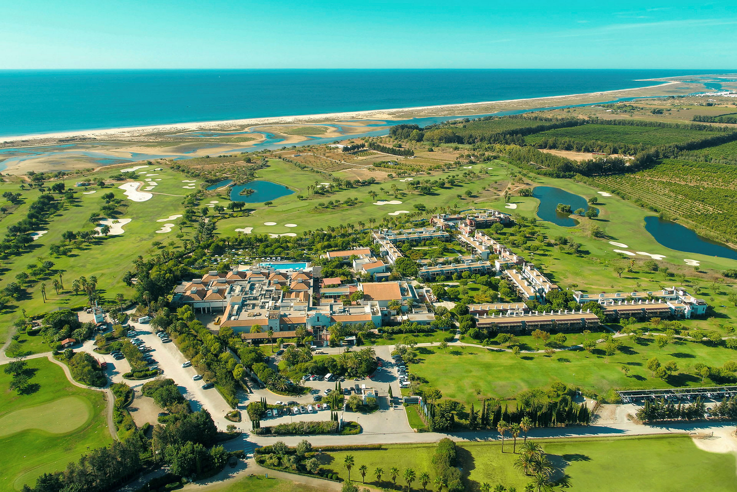 Luftfoto af et feriekompleks omgivet af grønne områder, golfbaner og nærhed til havet. (AI-genereret tekst)