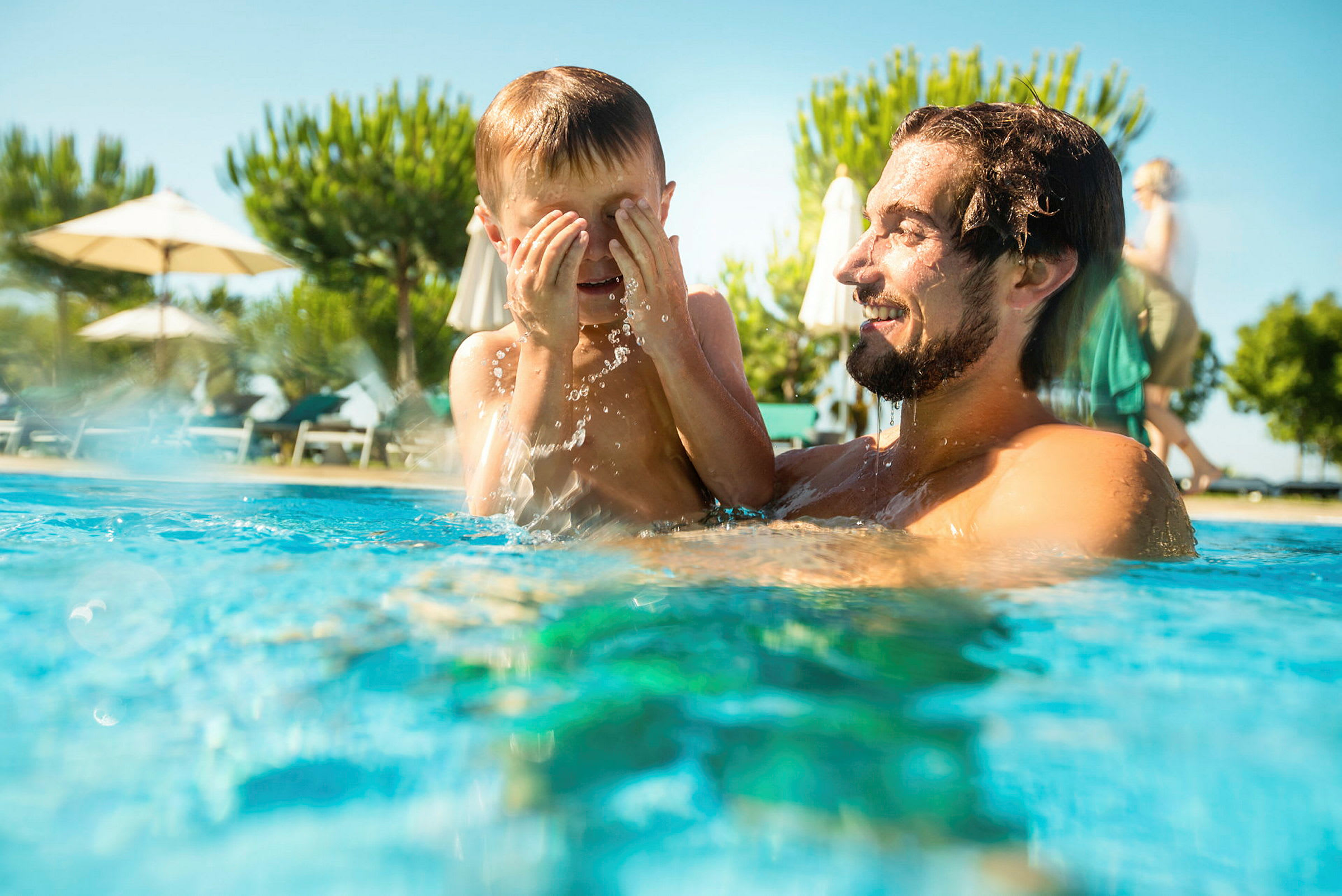 Personer nyder en solrig dag i en pool med parasoller og træer i baggrunden. (AI-genereret tekst)