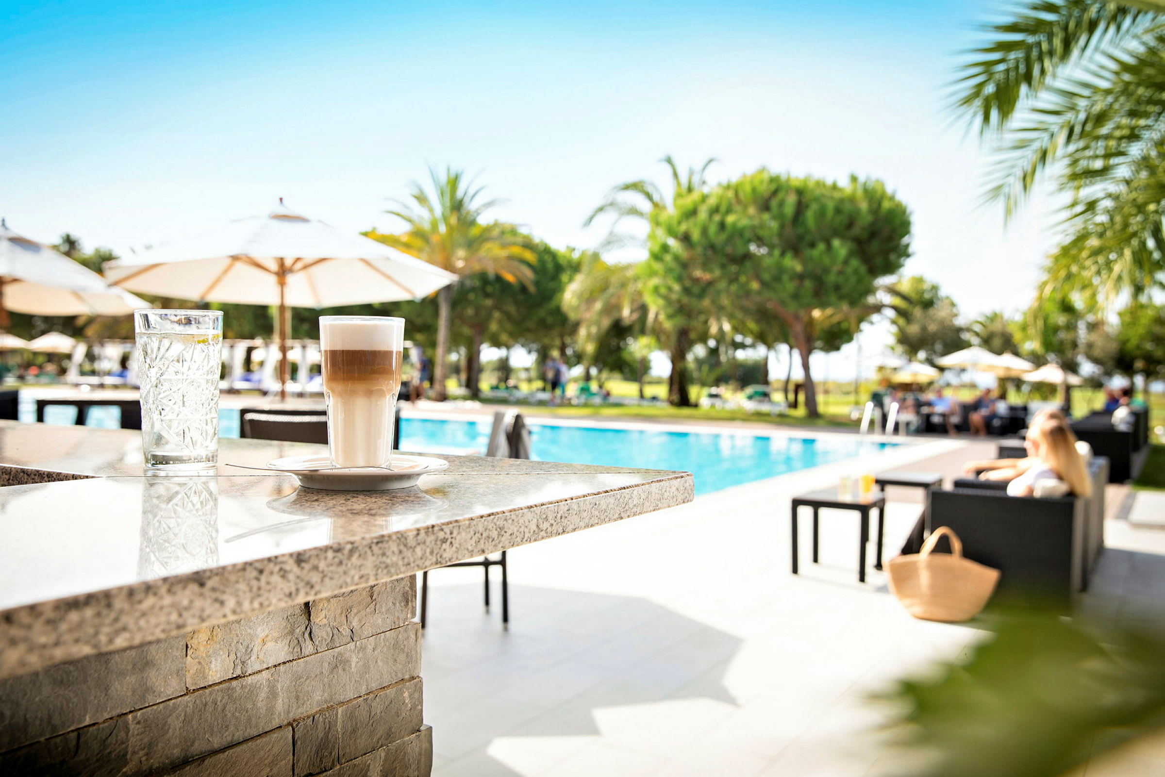 Et glas vand og en latte på en stenbar ved en pool med omkringliggende liggestole og parasoller. (AI-genereret tekst)