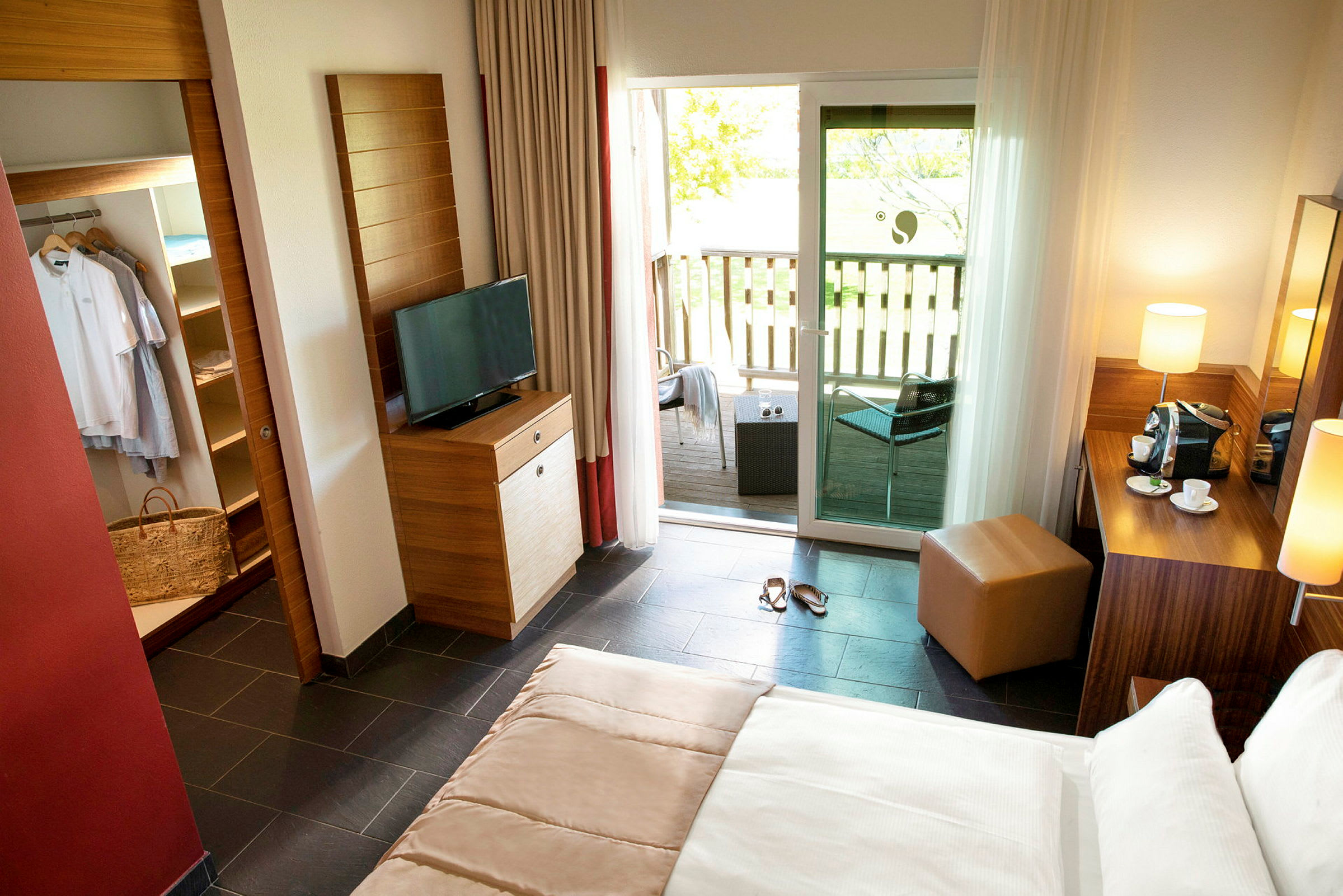 Et lyst hotelværelse med seng, TV, et garderobeskab og en balkon med siddeområde udenfor. (AI-genereret tekst)