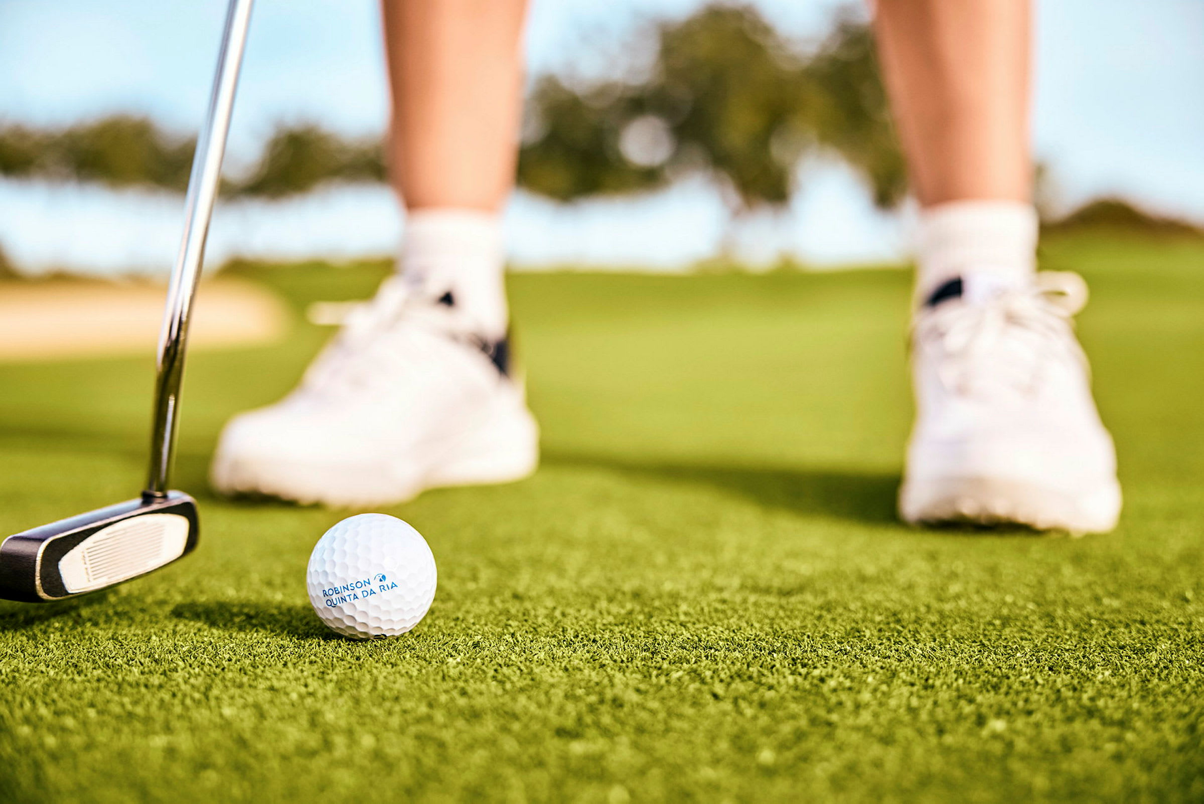 En golfbold og en putter på en grøn græsplæne, med en persons fødder iført hvide sportssko i baggrunden. (AI-genereret tekst)