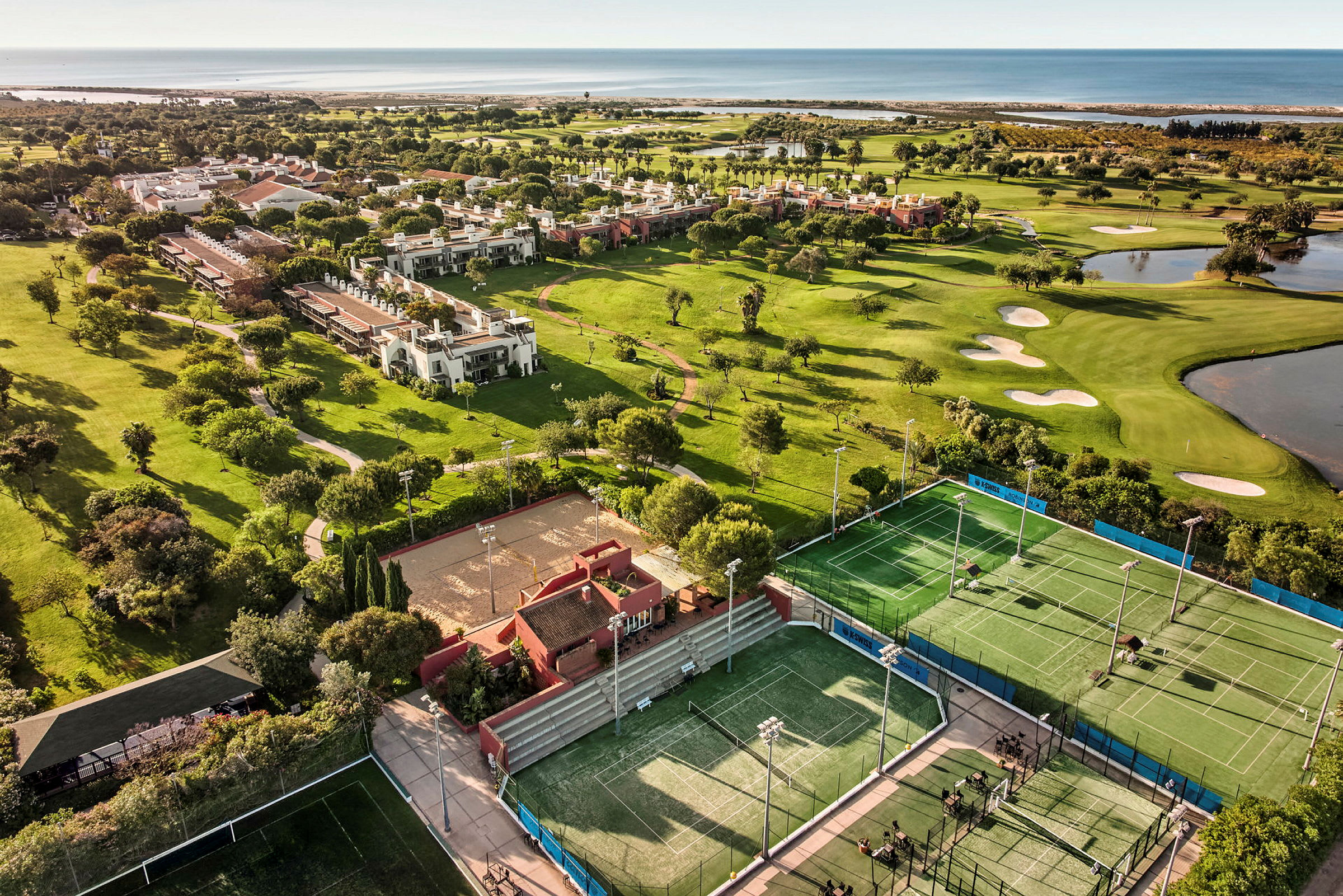Luftfoto af et resort med tennisbaner, en golfbane og nærliggende bygninger omgivet af grønne områder og tæt på havet. (AI-genereret tekst)