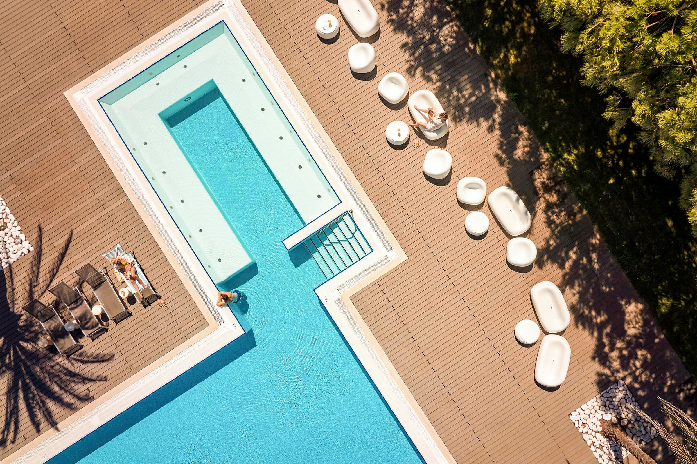 Luftfoto af en pool omgivet af et dæk med liggestole og siddepladser samt personer, der slapper af. (AI-genereret tekst)