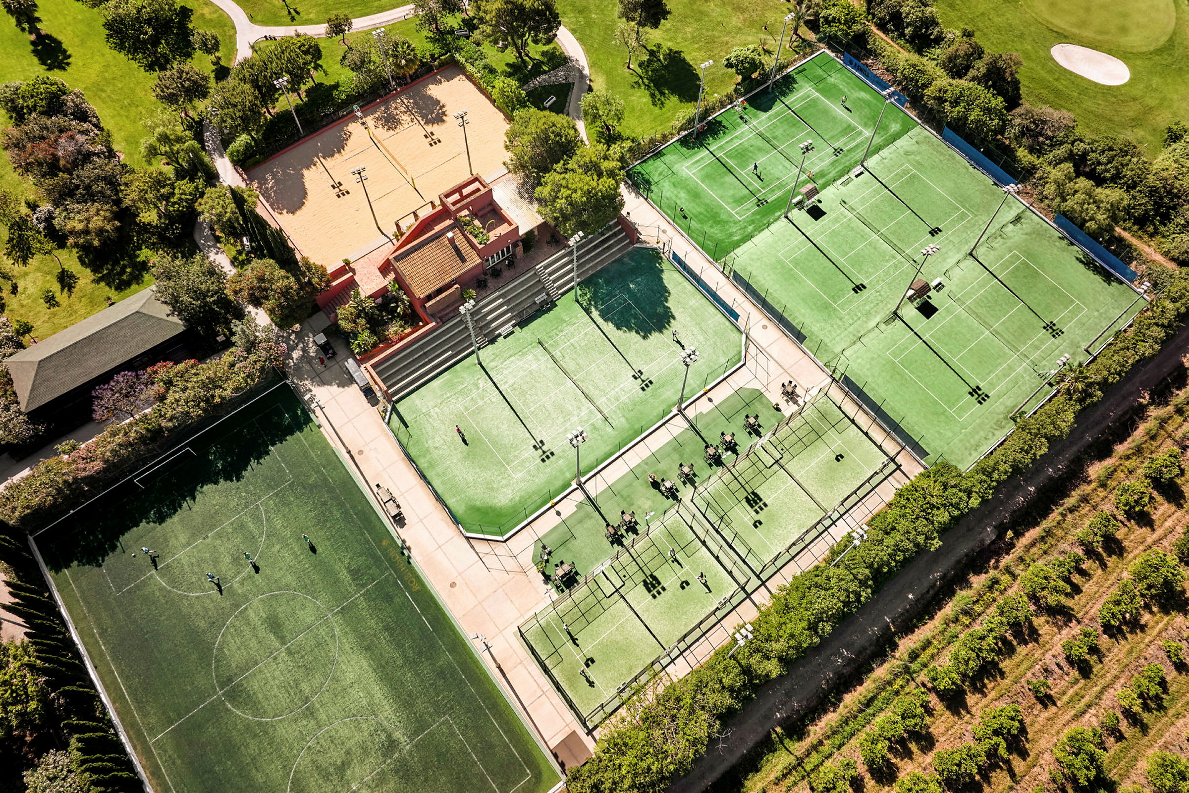 Luftfoto af et sportsanlæg med tennisbaner, padelbaner, en fodboldbane og et sandområde, omgivet af grønne områder. (AI-genereret tekst)