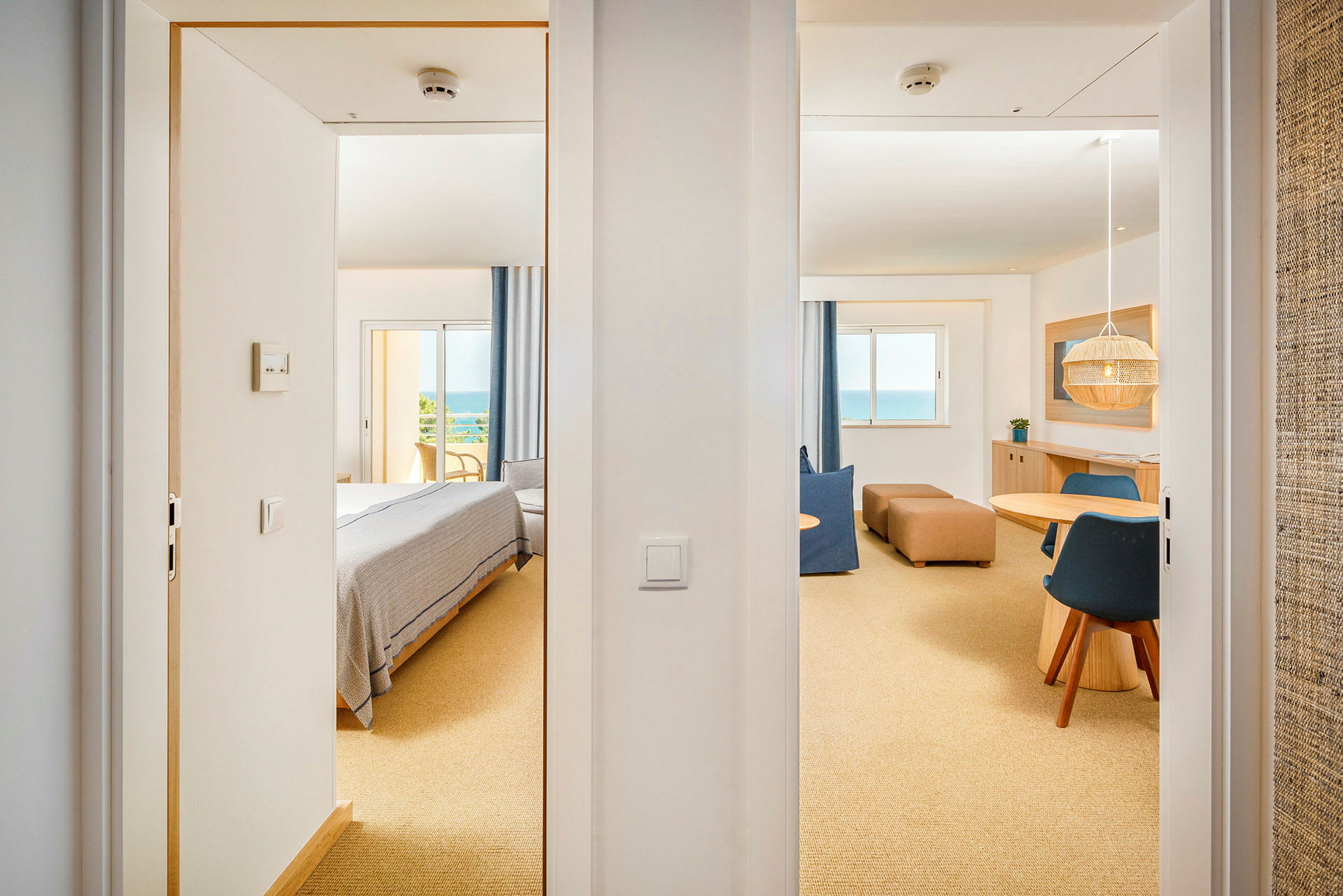 Udsigt gennem to døråbninger til et lyst hotelværelse med soveområde og siddegruppe samt et stort vindue med havudsigt. (AI-genereret tekst)
