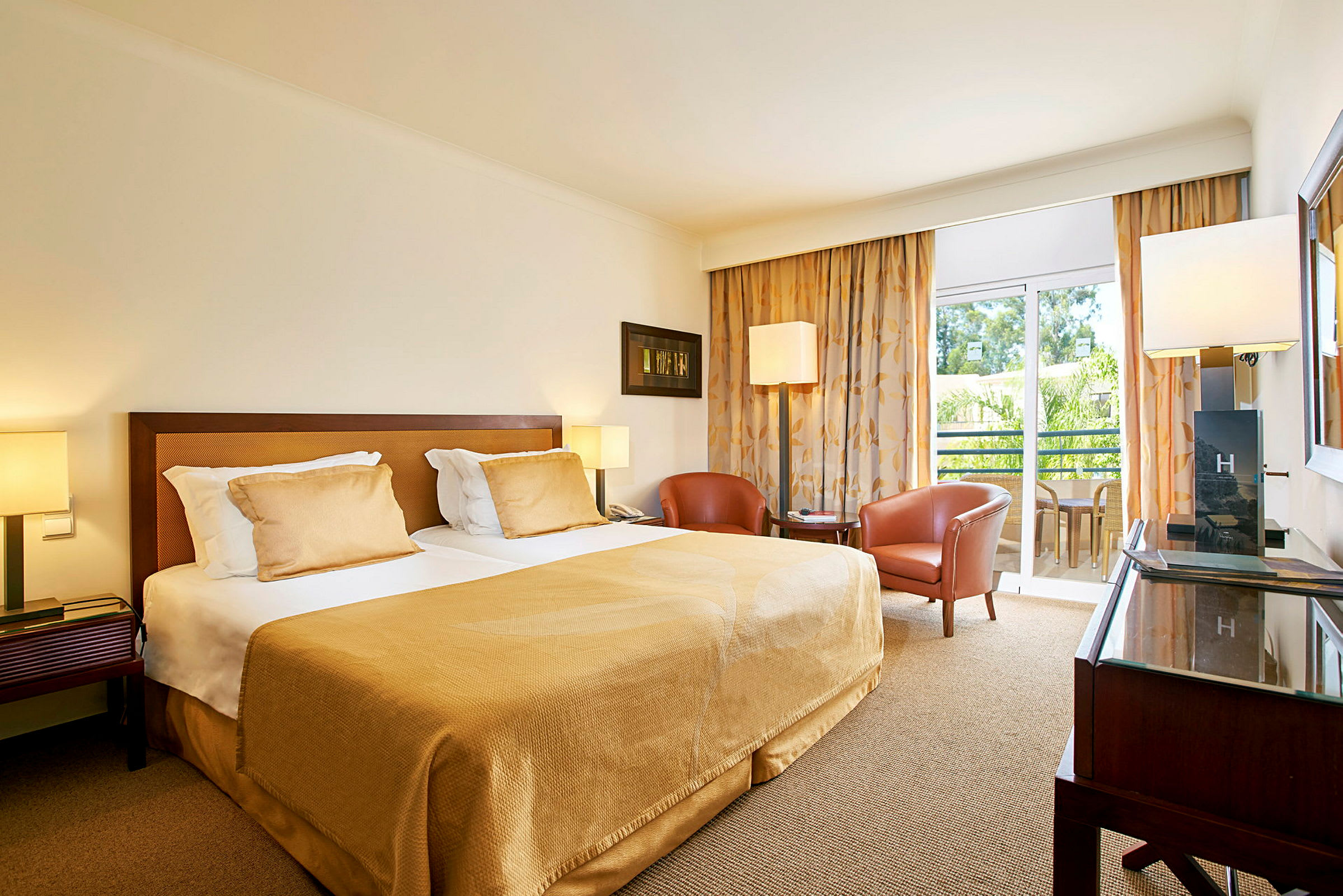 Et hotelværelse med en dobbeltseng, to lænestole og et bord. På sengen er lyse puder og tæppe. Et vindue og en balkon er synlig i baggrunden. (AI-genereret tekst)