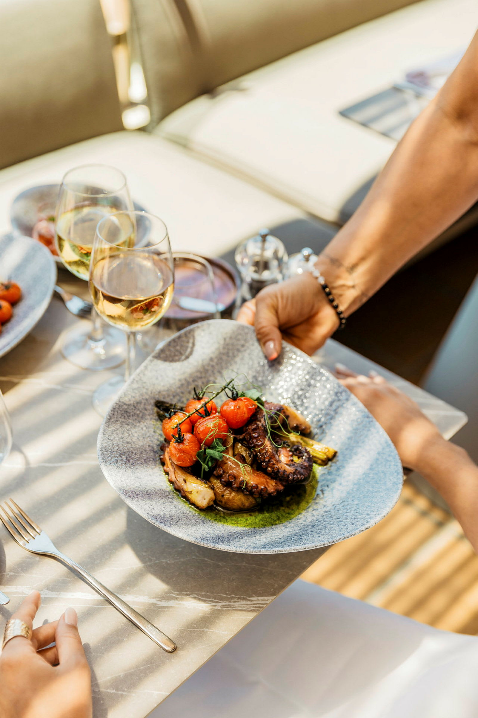Billedet viser en tallerken med en delikat ret bestående af grillet blæksprutte, tomater og grøntsager, serveret på et bord med vinglas. (AI-genereret tekst)