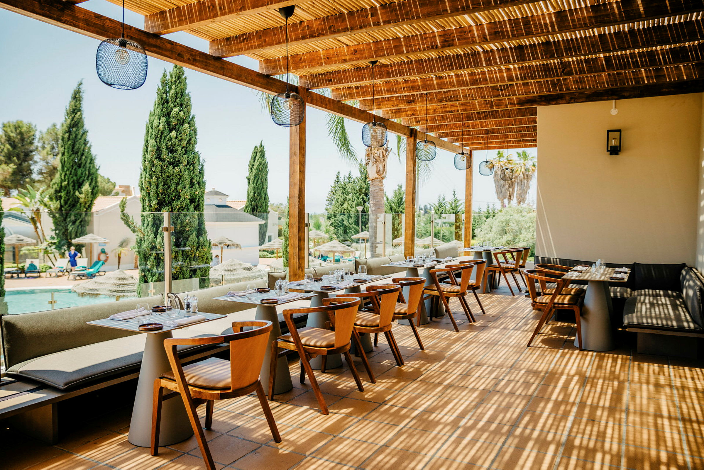 Restaurant udendørs terrasse med borde og stole, med udsigt over en pool og træer. (AI-genereret tekst)