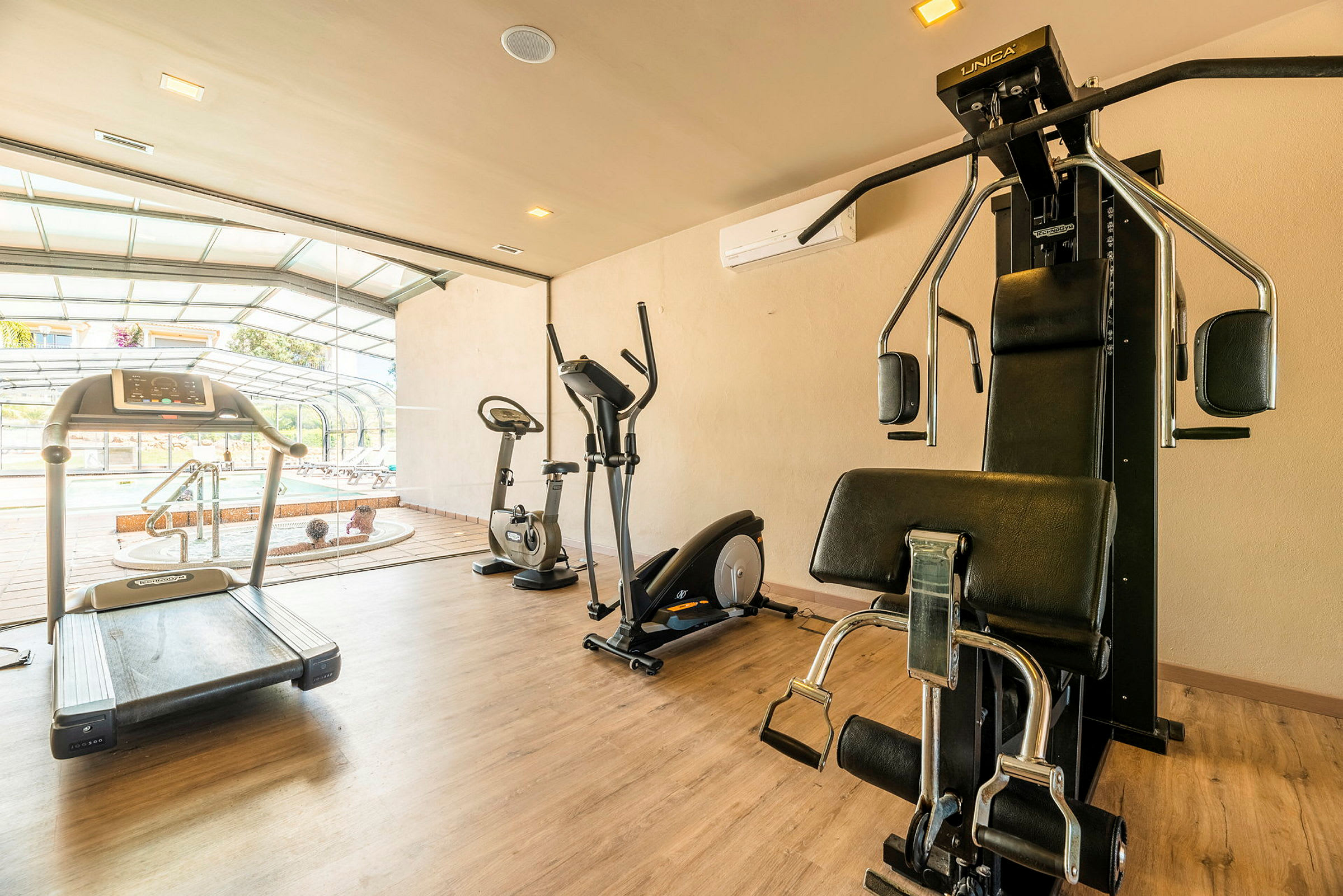 Billede af et fitnesscenter med løbebånd, motionscykler og styrketræningsudstyr. I baggrunden ses et boblebad og en terrasse. (AI-genereret tekst)