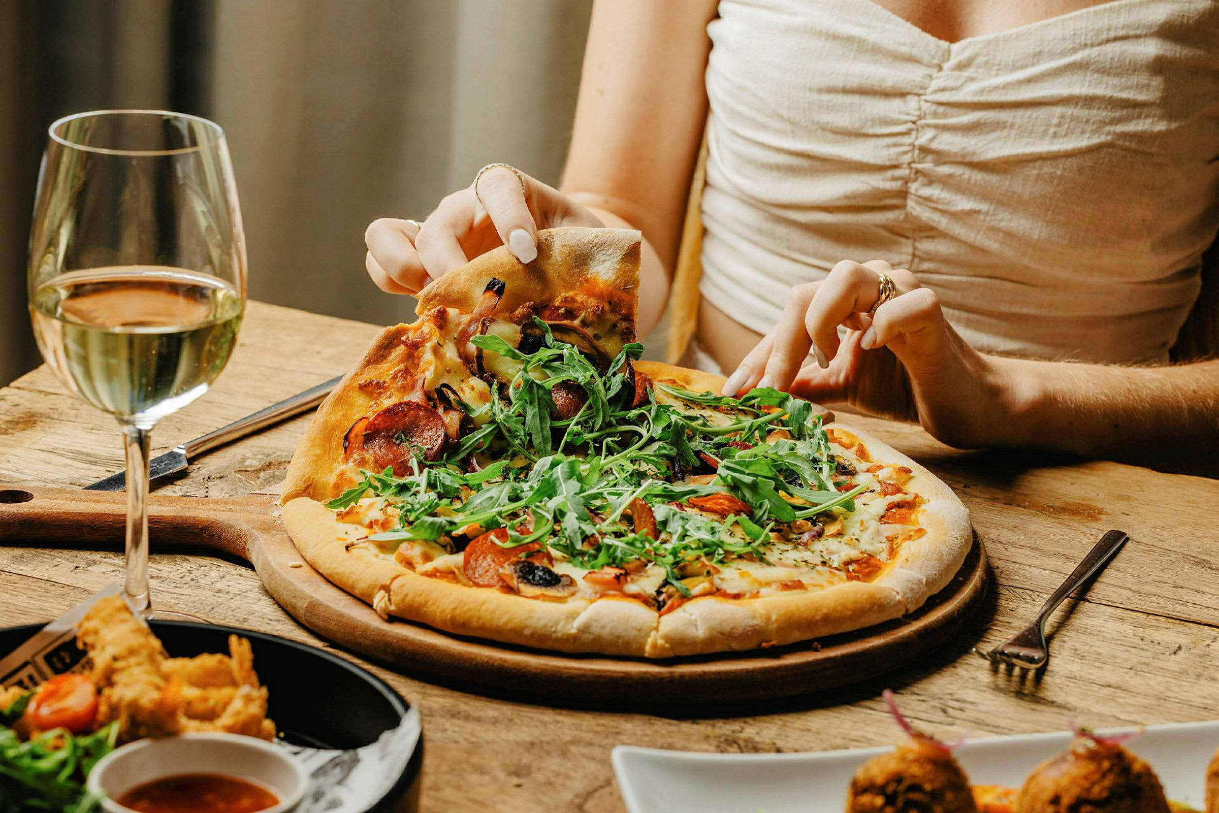 Nærbillede af en person, der tager et stykke pizza fra en tallerken med et glas hvidvin ved siden af. (AI-genereret tekst)