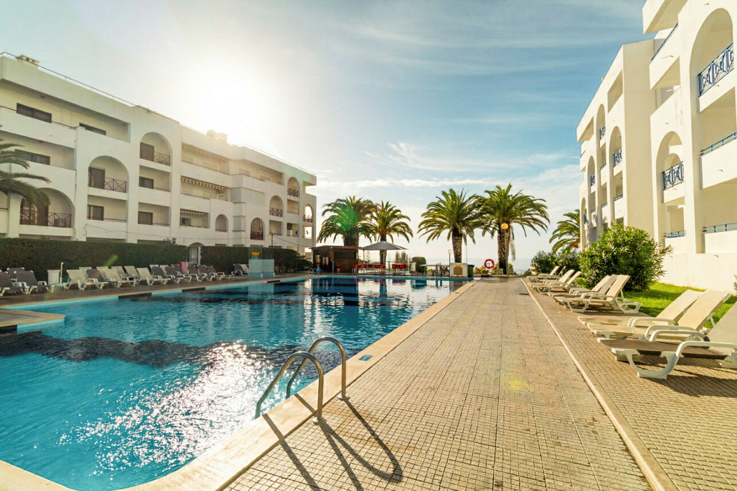 Hotelpool omgivet af liggestole og palmer med en bygning i baggrunden. (AI-genereret tekst)