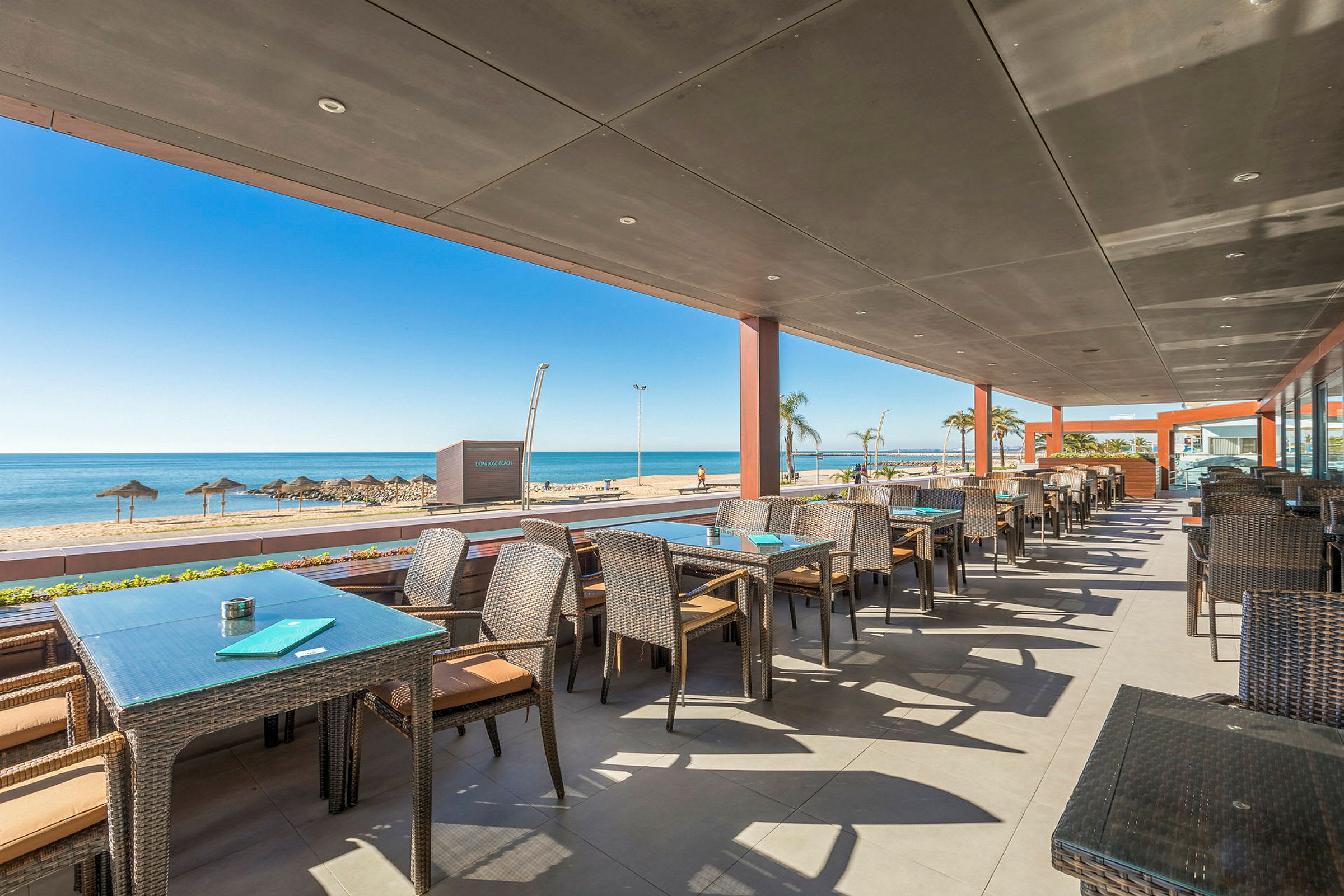 Restaurant med udendørs servering og udsigt til stranden og havet. (AI-genereret tekst)